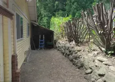 Narrow gravel side yard beside a house, with a ladder and trimmed bushes along a stone border.