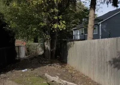 Narrow dirt path beside a wooden fence, trees, and a gray house in a shaded yard.