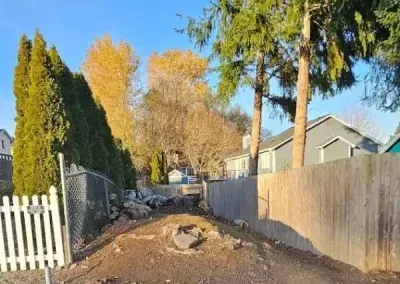 Narrow dirt alley between a white fence and gray wall, with trees and houses in autumn light