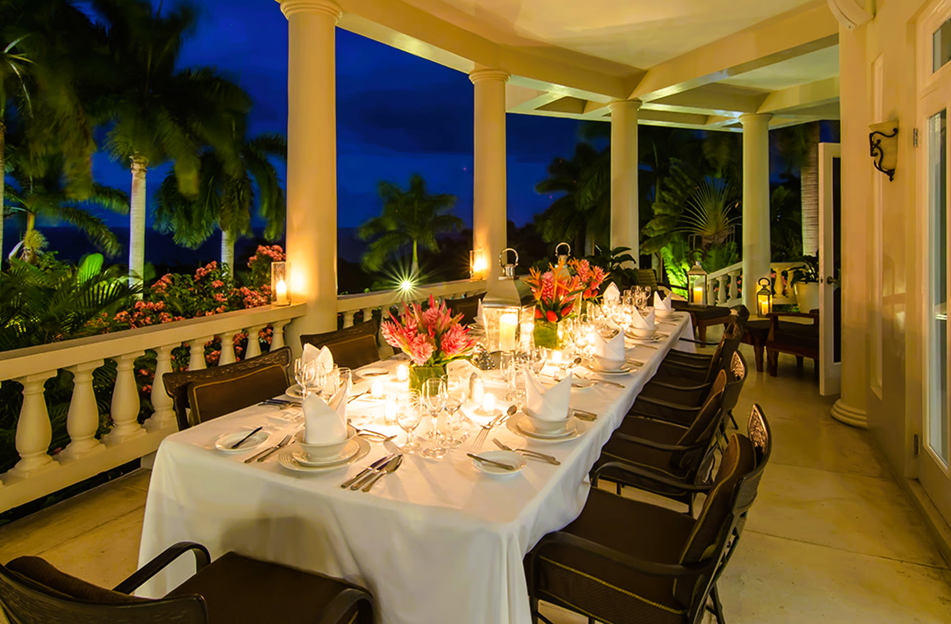 Elegant outdoor dinner table set for many guests, illuminated by candles, with a tropical backdrop.