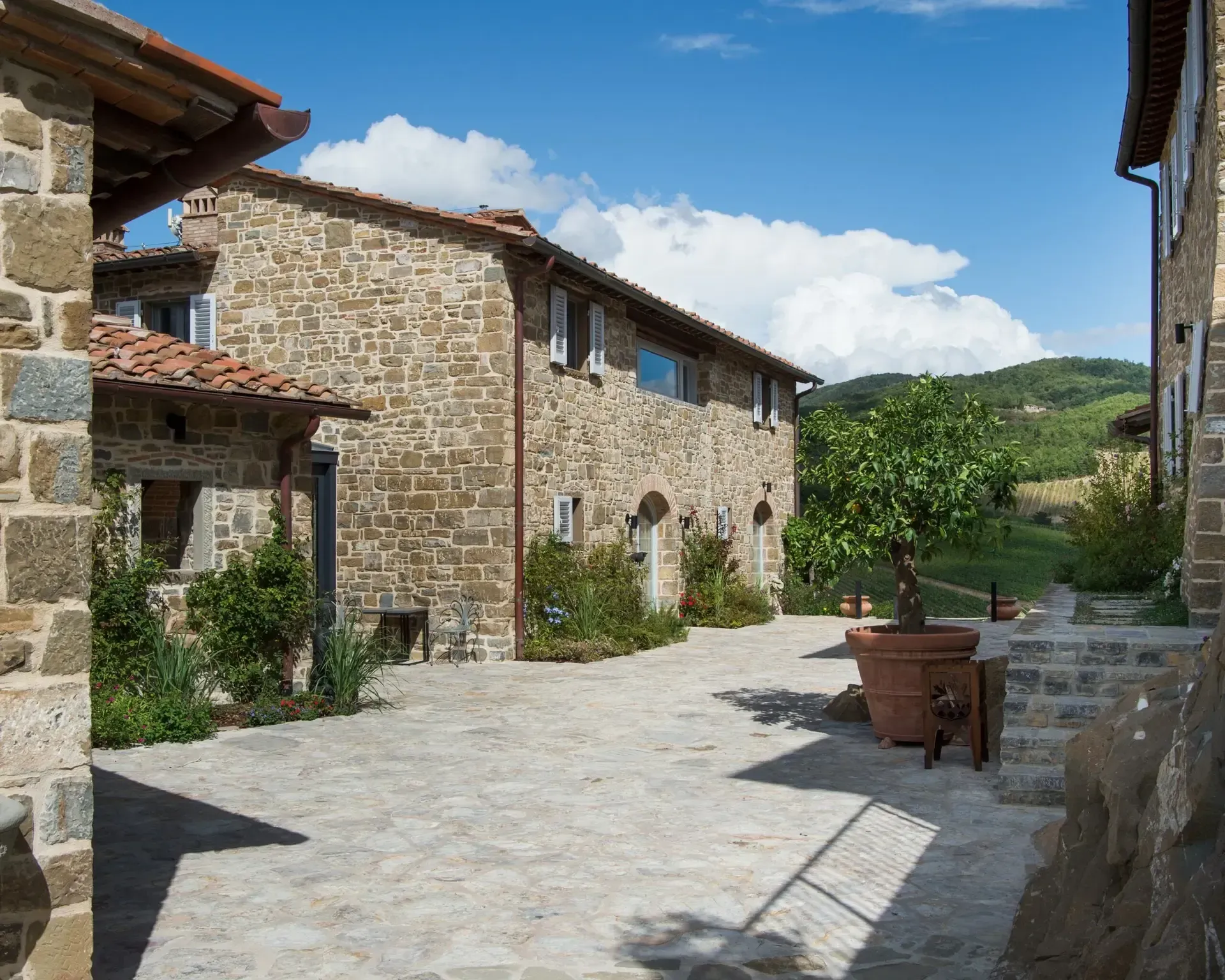 Stone buildings in a sunlit courtyard, with a potted tree and a hillside in the background under a blue sky.