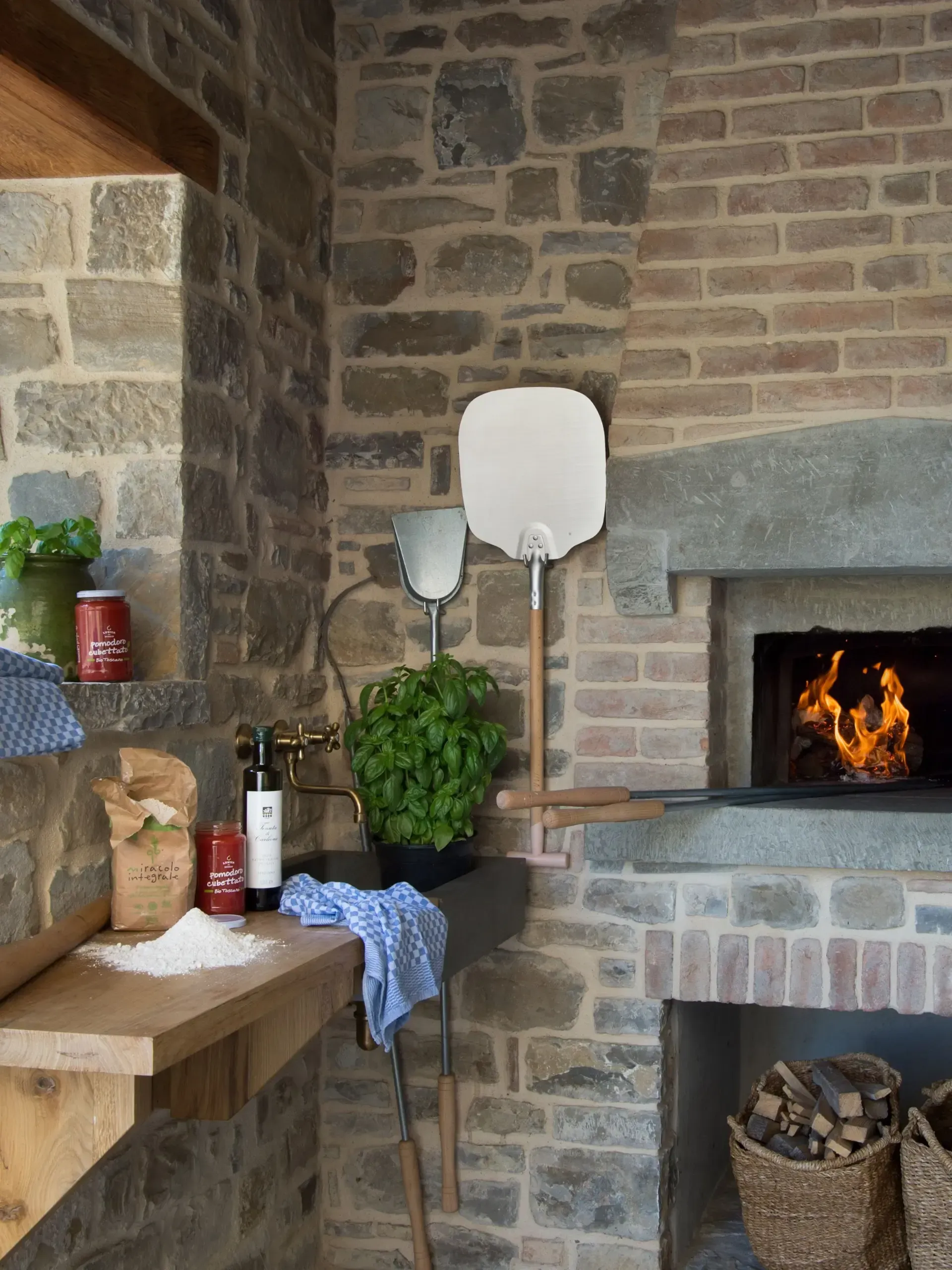Brick pizza oven with tools, herbs, and prep area in a stone-walled outdoor kitchen.