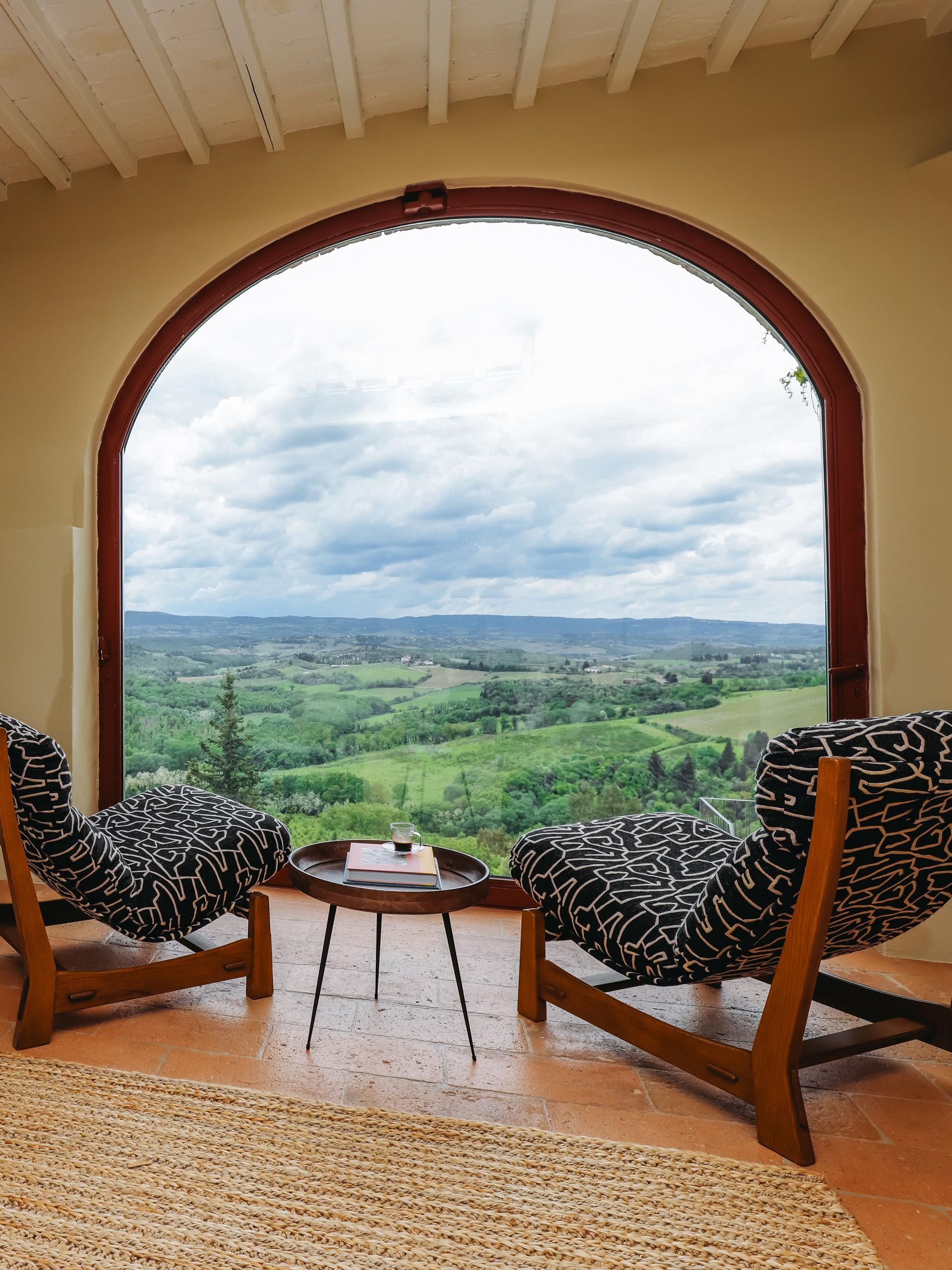 Two patterned chairs face a small table by an arched window with a scenic countryside view.
