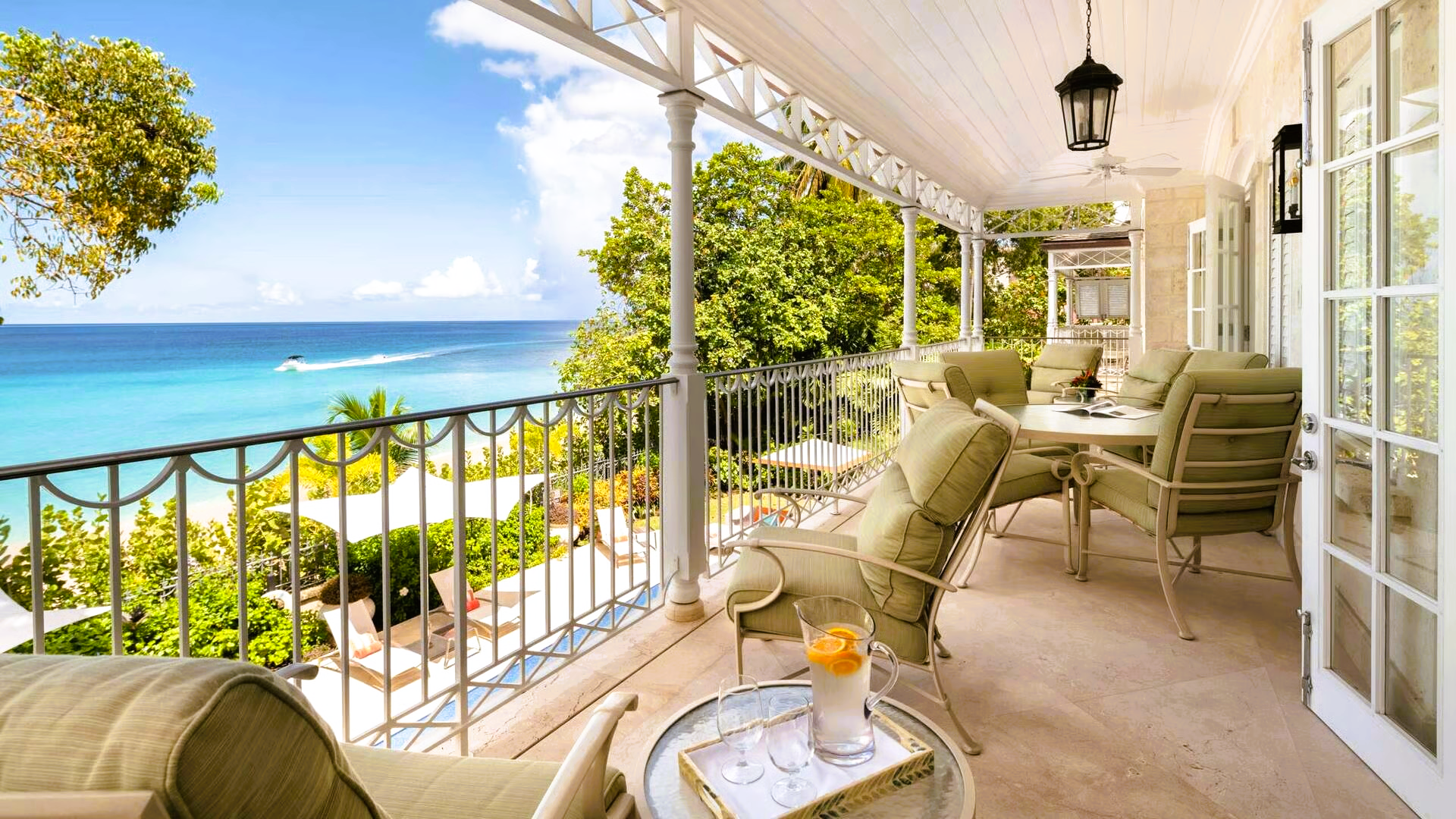 Balcony overlooking turquoise ocean; lounge chairs, table, and trees are present.
