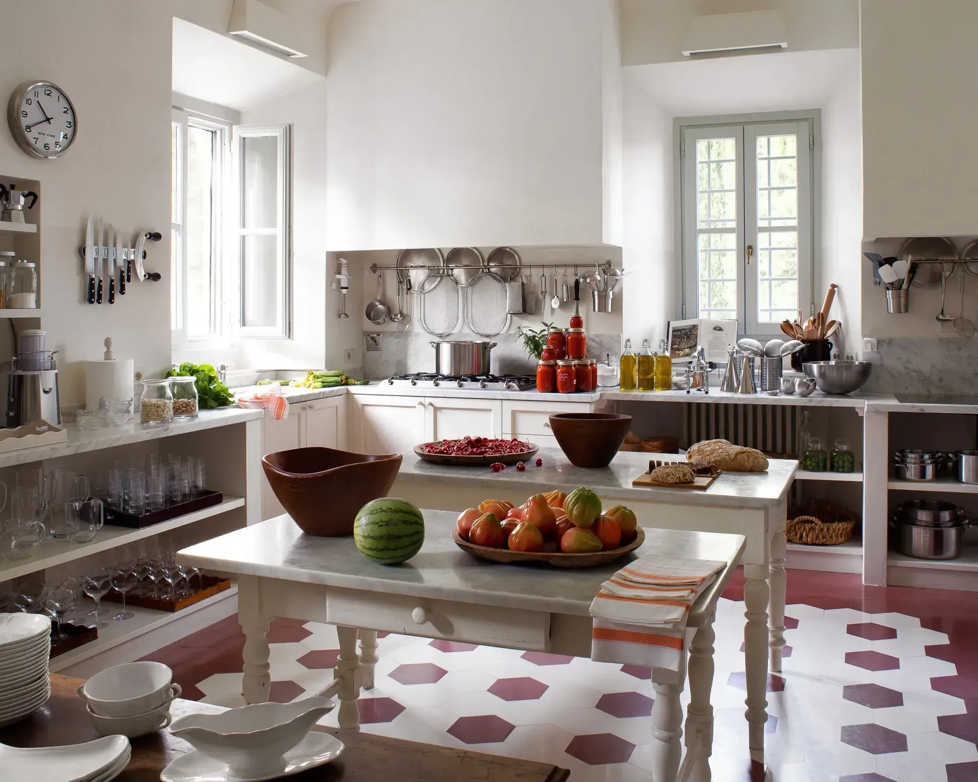 Bright kitchen with white cabinets, two tables, fresh produce, and red tile floor.