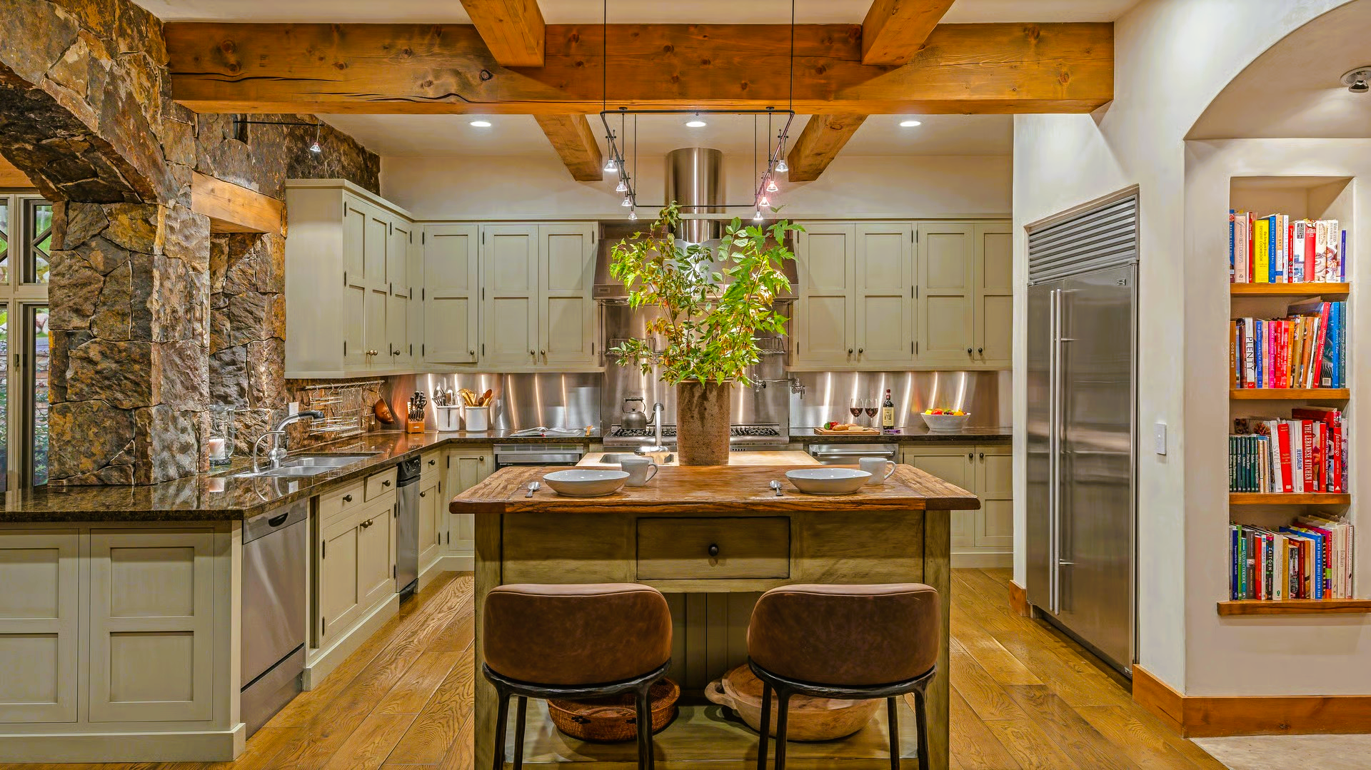 Rustic kitchen with stone wall, light green cabinets, wooden beams, island with stools, and built-in bookshelves.