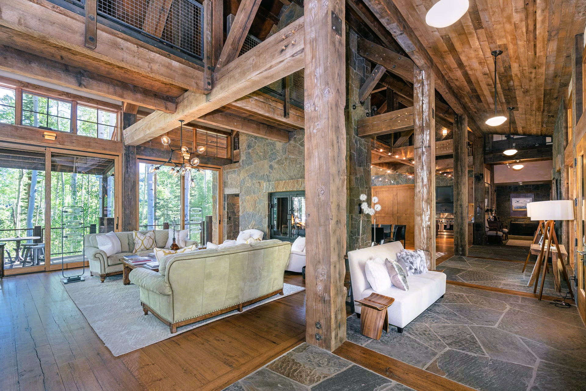 Rustic, open-concept living room with exposed wooden beams, stone wall, and multiple seating areas, overlooking forest.