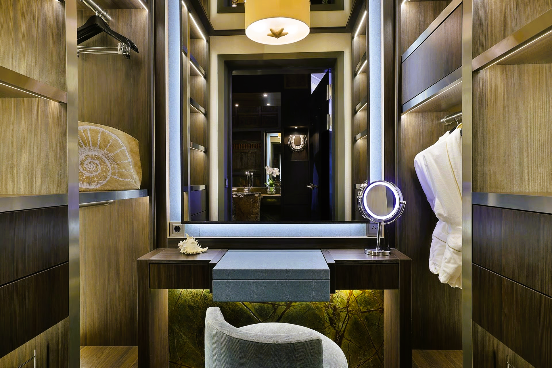 Luxurious walk-in closet with lighted vanity, shelves, and an upholstered chair. Neutral tones.