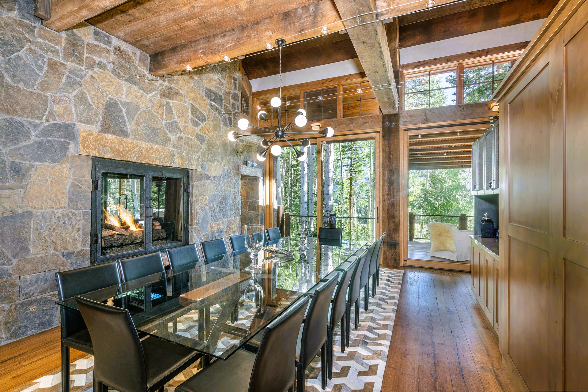 Dining room with long glass table, stone fireplace, and wooden beams.