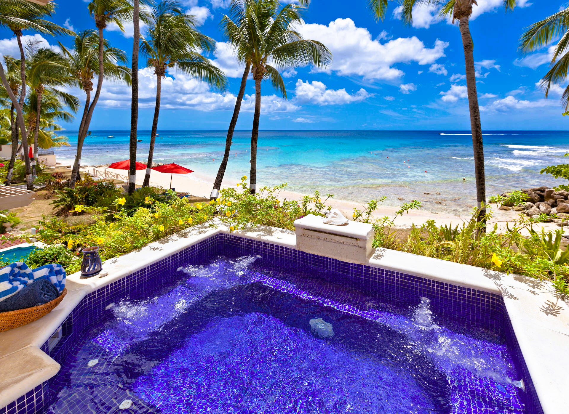 Hot tub overlooking a white sand beach, turquoise water, palm trees, and blue sky.