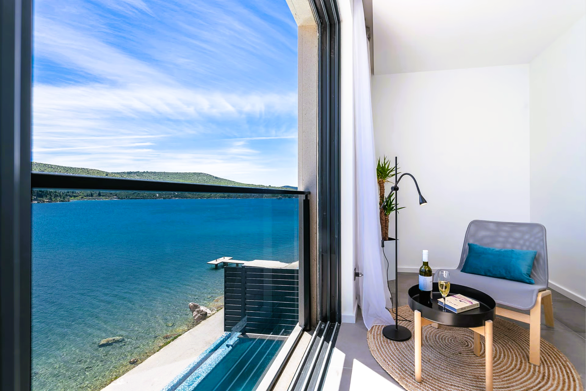 Room with water view: chair, table with wine, sheer curtain, and large window overlooking a blue lake.