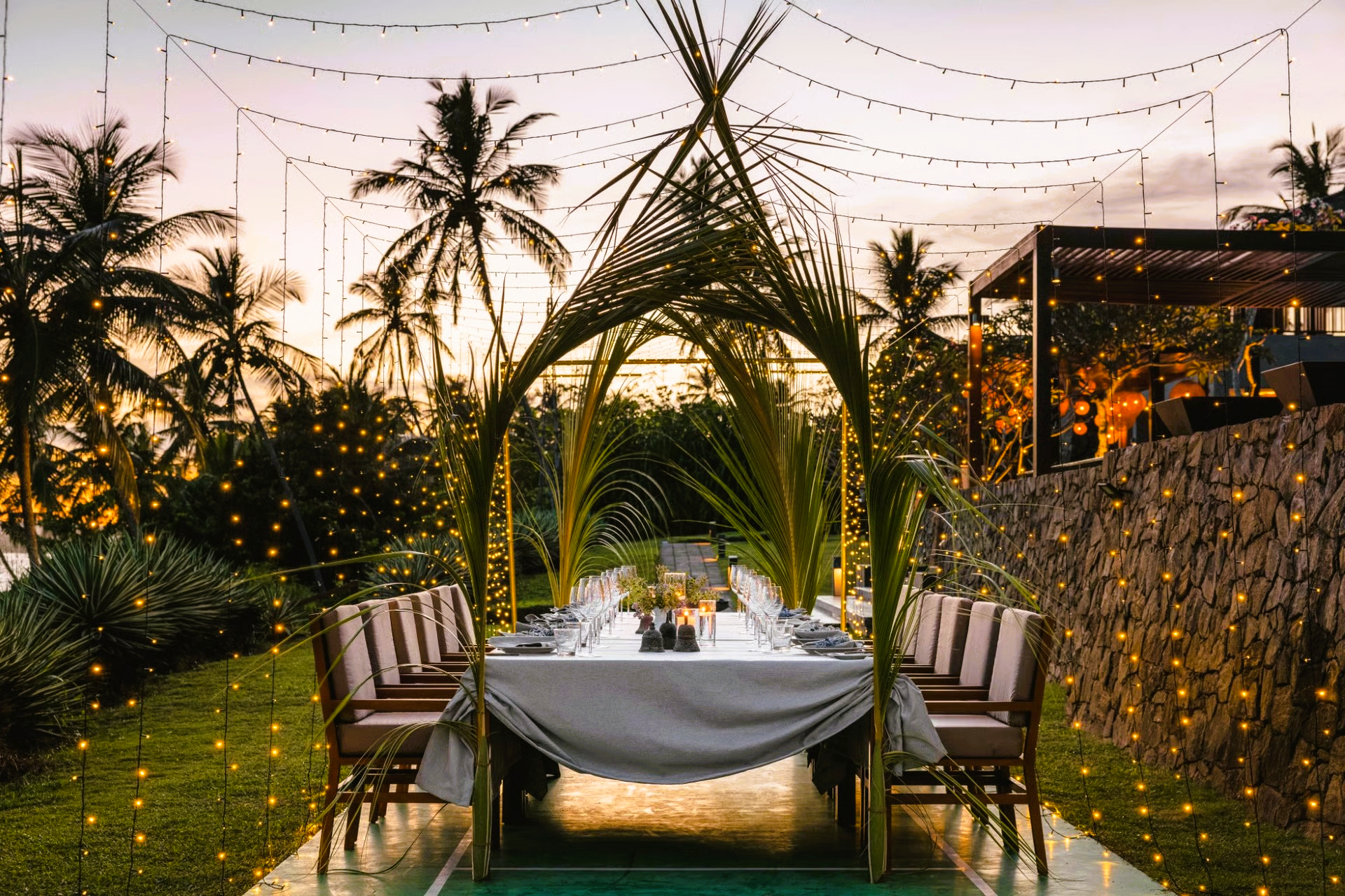 Outdoor dining table set for a celebration, lit by string lights and palm frond arches at sunset.
