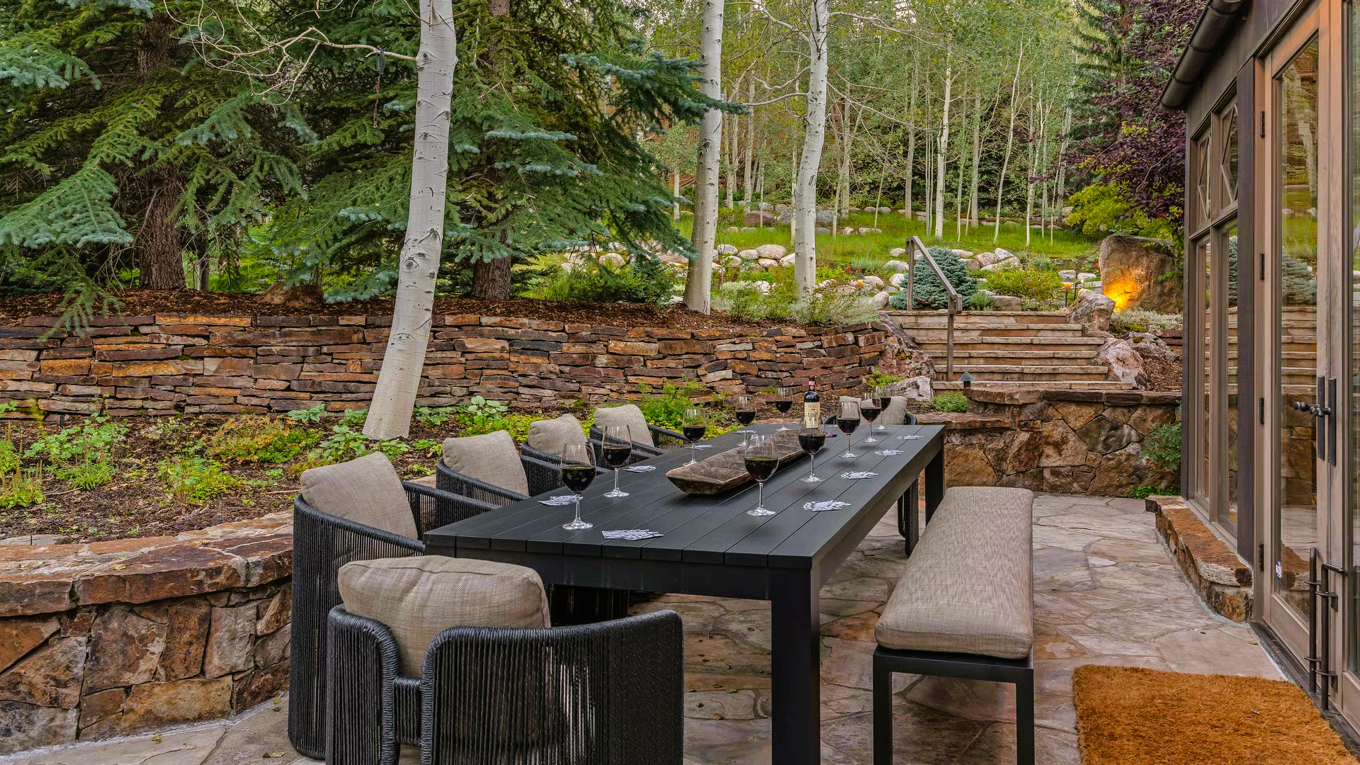 Outdoor dining area with a long black table and chairs, stone walls, and trees.
