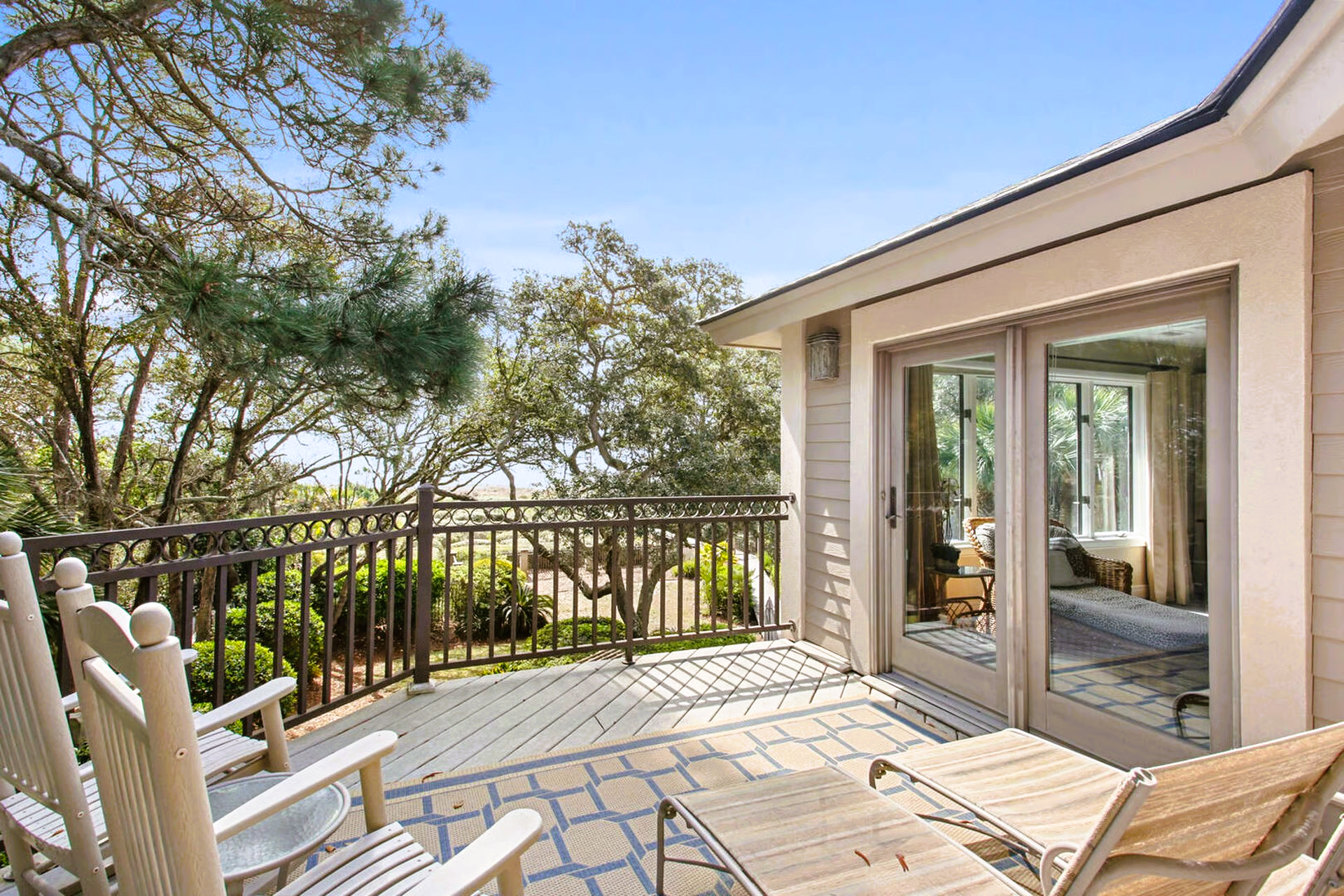Balcony with rocking chairs, sun lounger, and trees, with glass doors leading to a room.