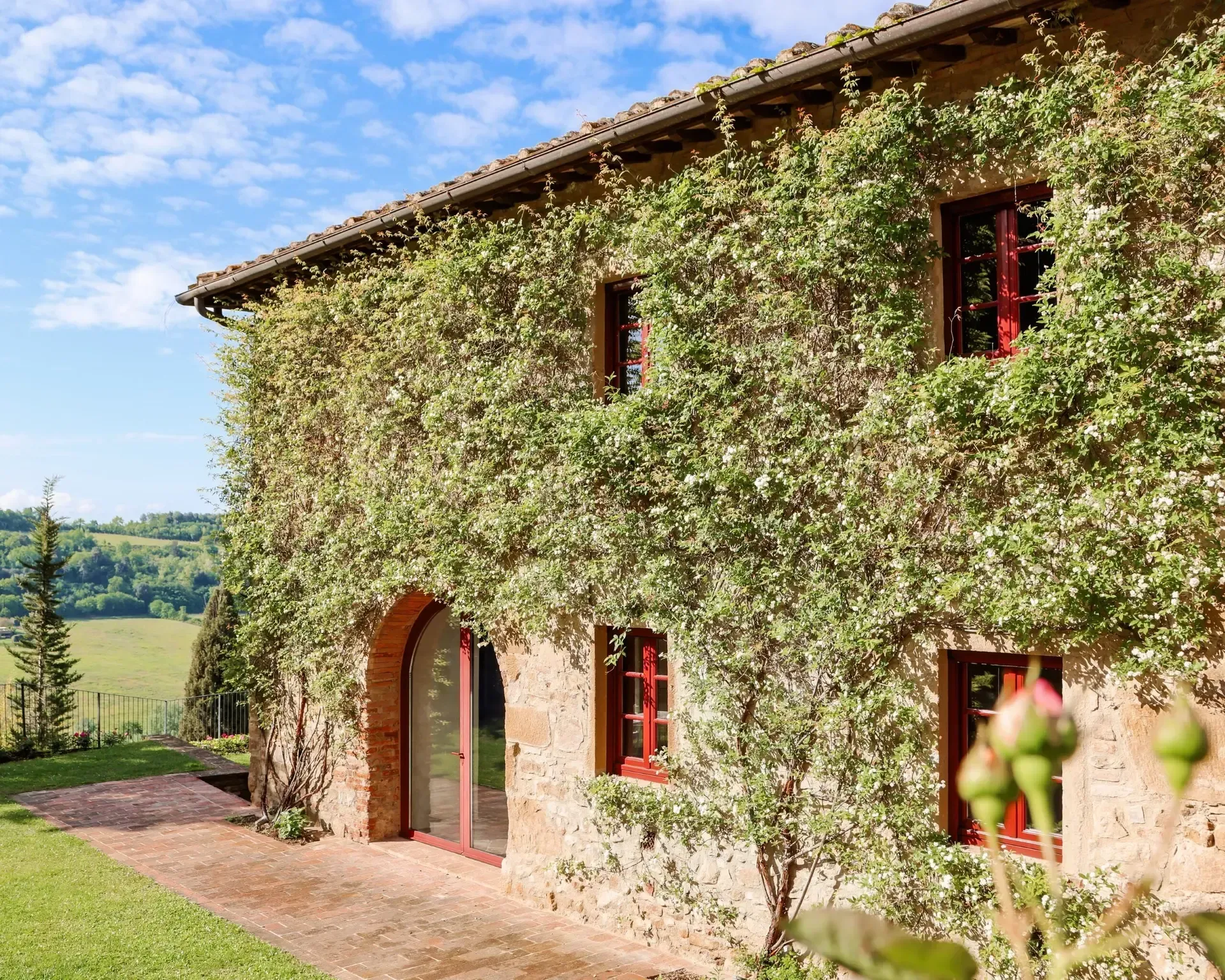 Stone building covered in green vines with red-trimmed windows and doors, set against a blue sky and rolling hills.