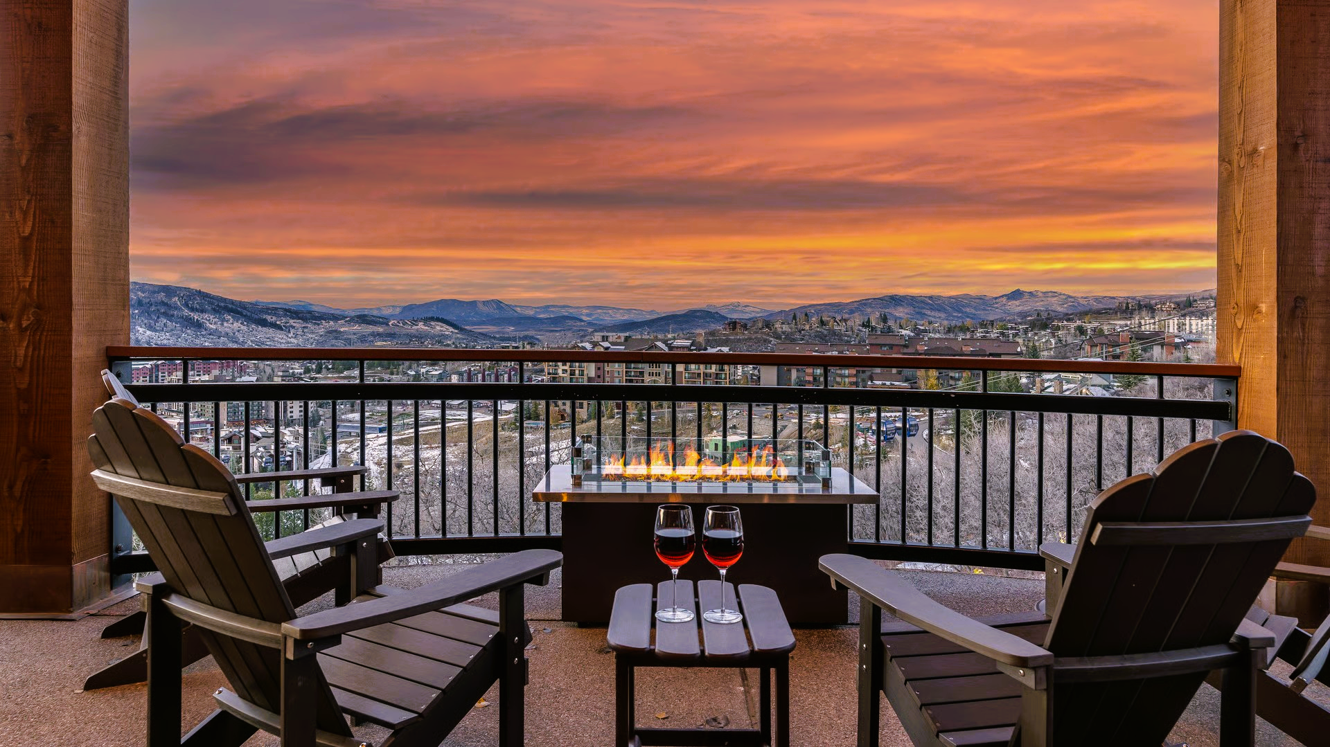 Balcony with two chairs, a fire pit, and drinks overlooking a cityscape at sunset.