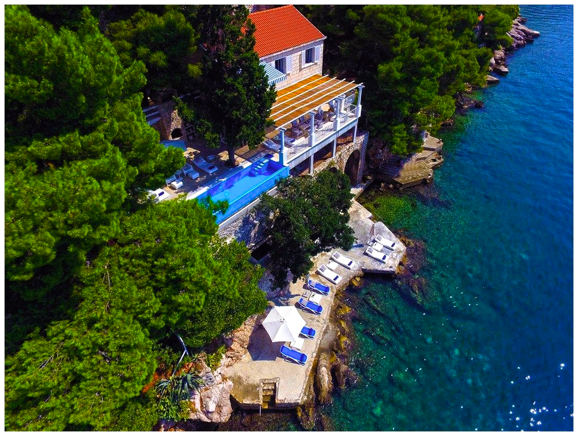 Coastal villa with pool, trees, and seating area over blue sea.