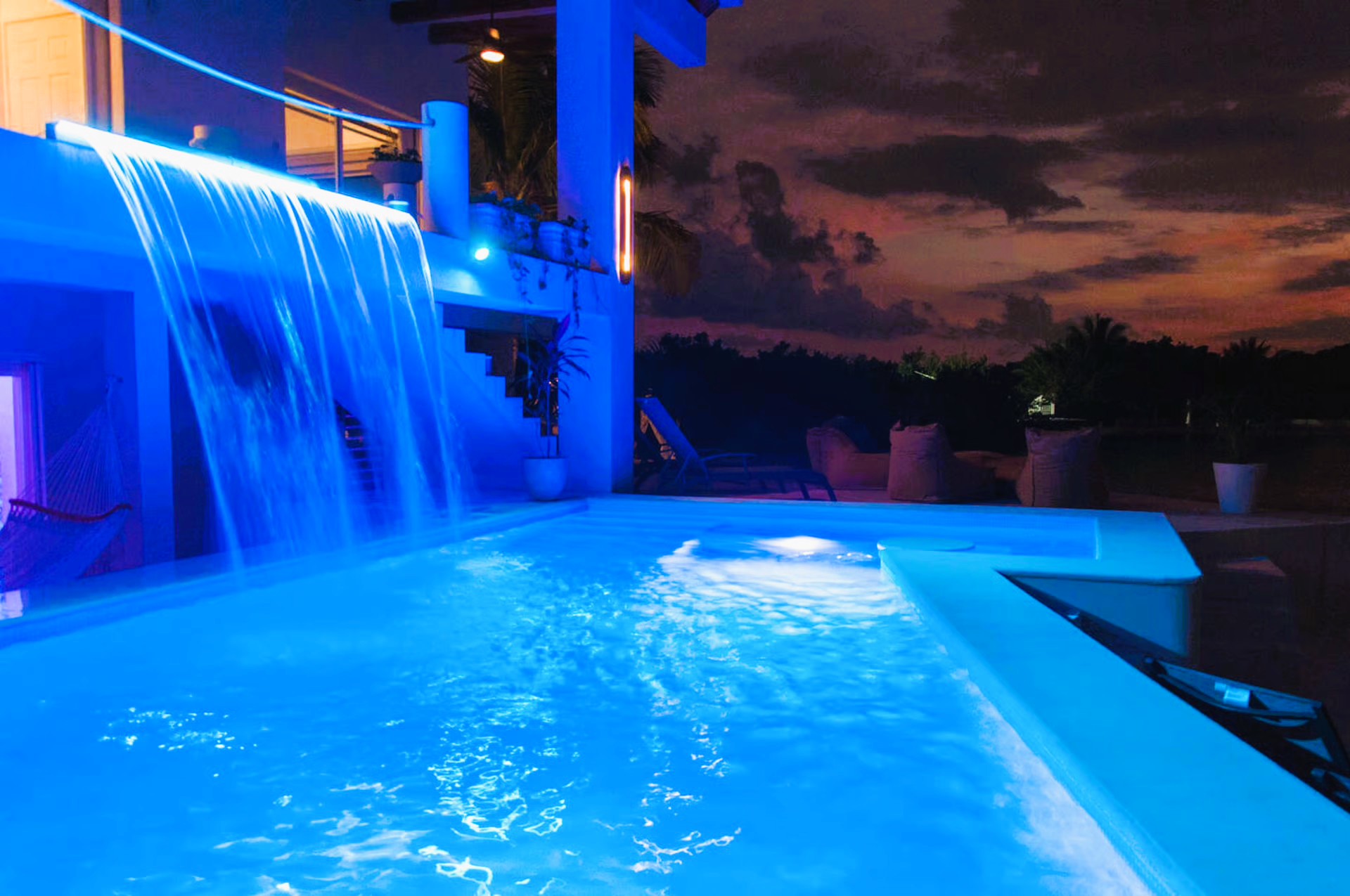 Pool illuminated blue at night; waterfall cascading; white building and pool surround.