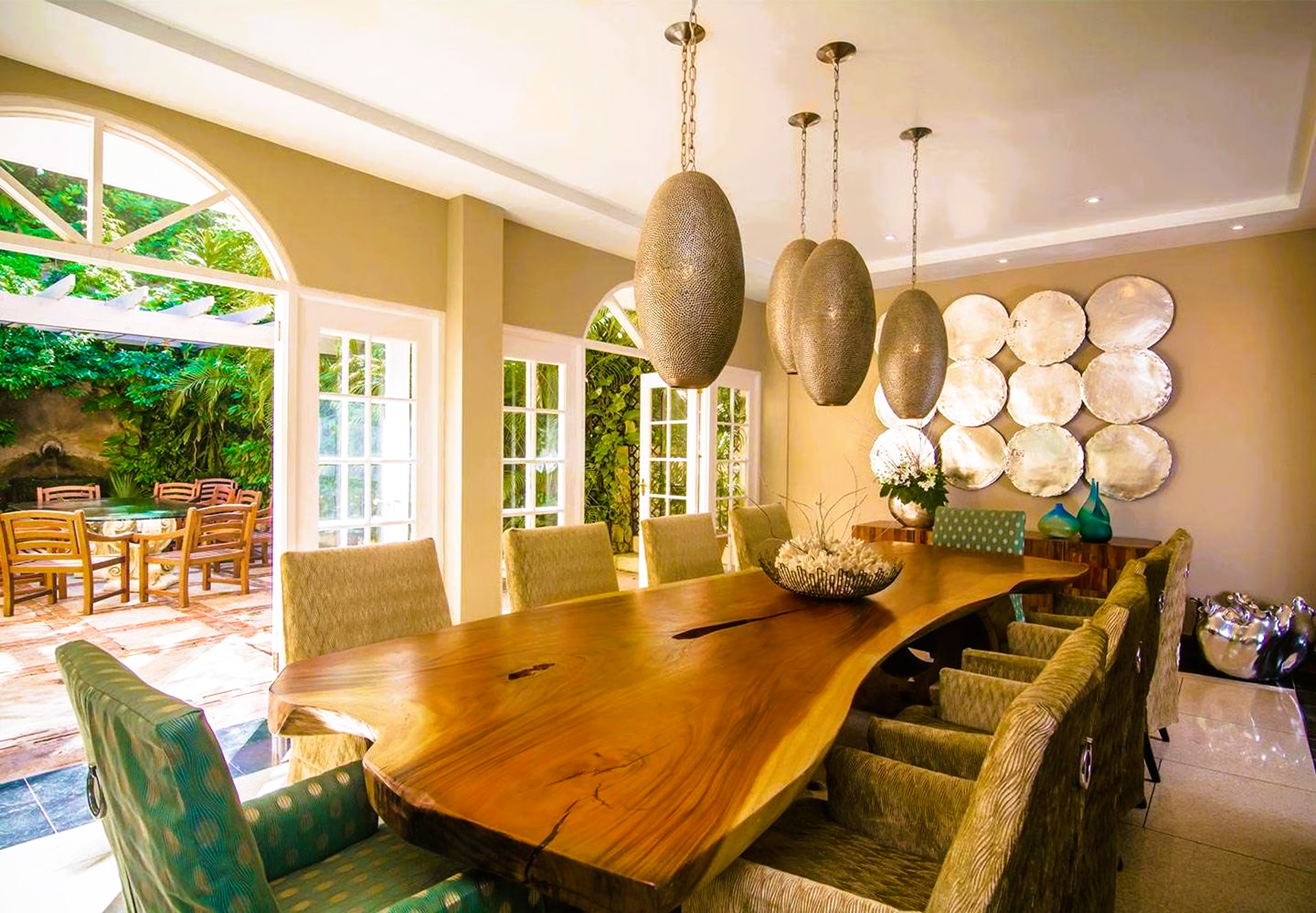Dining room with a long wooden table, elegant chairs, and hanging lights. Doors open to a patio.