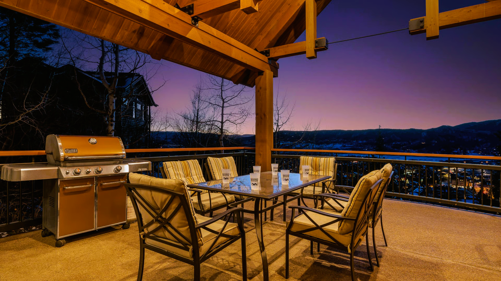 Patio with grill, table, chairs, and view of city lights at dusk.