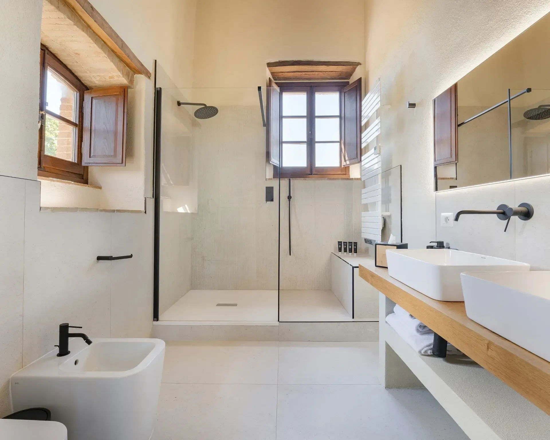 Modern white bathroom with shower, bidet, and floating double sink. Wooden window frames, black fixtures.