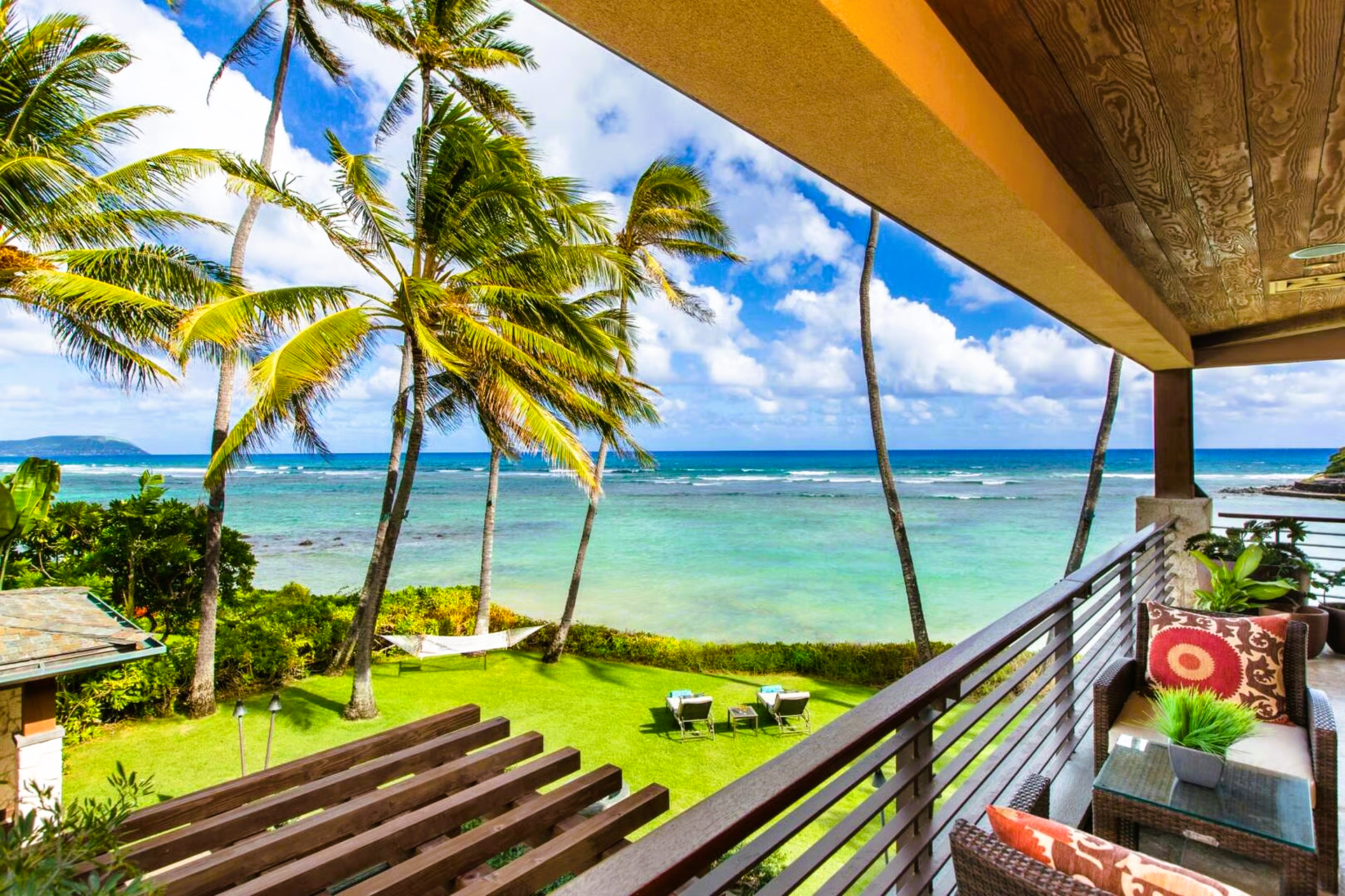 Ocean view from a balcony, green grass, palm trees, blue sky, chairs and a small table.