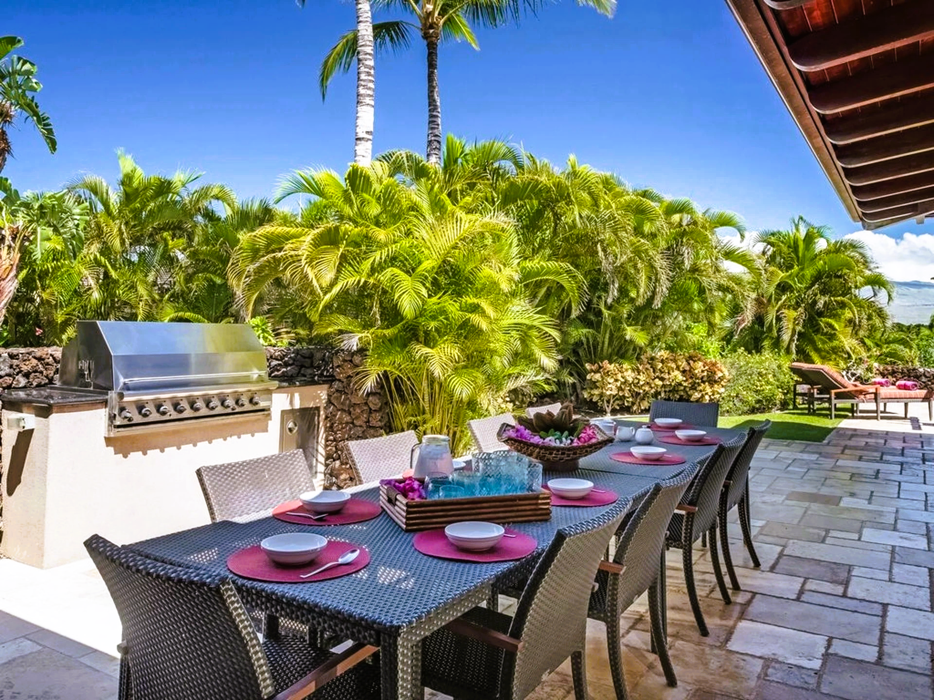 Outdoor dining table set for a meal, next to a grill and lush tropical foliage.
