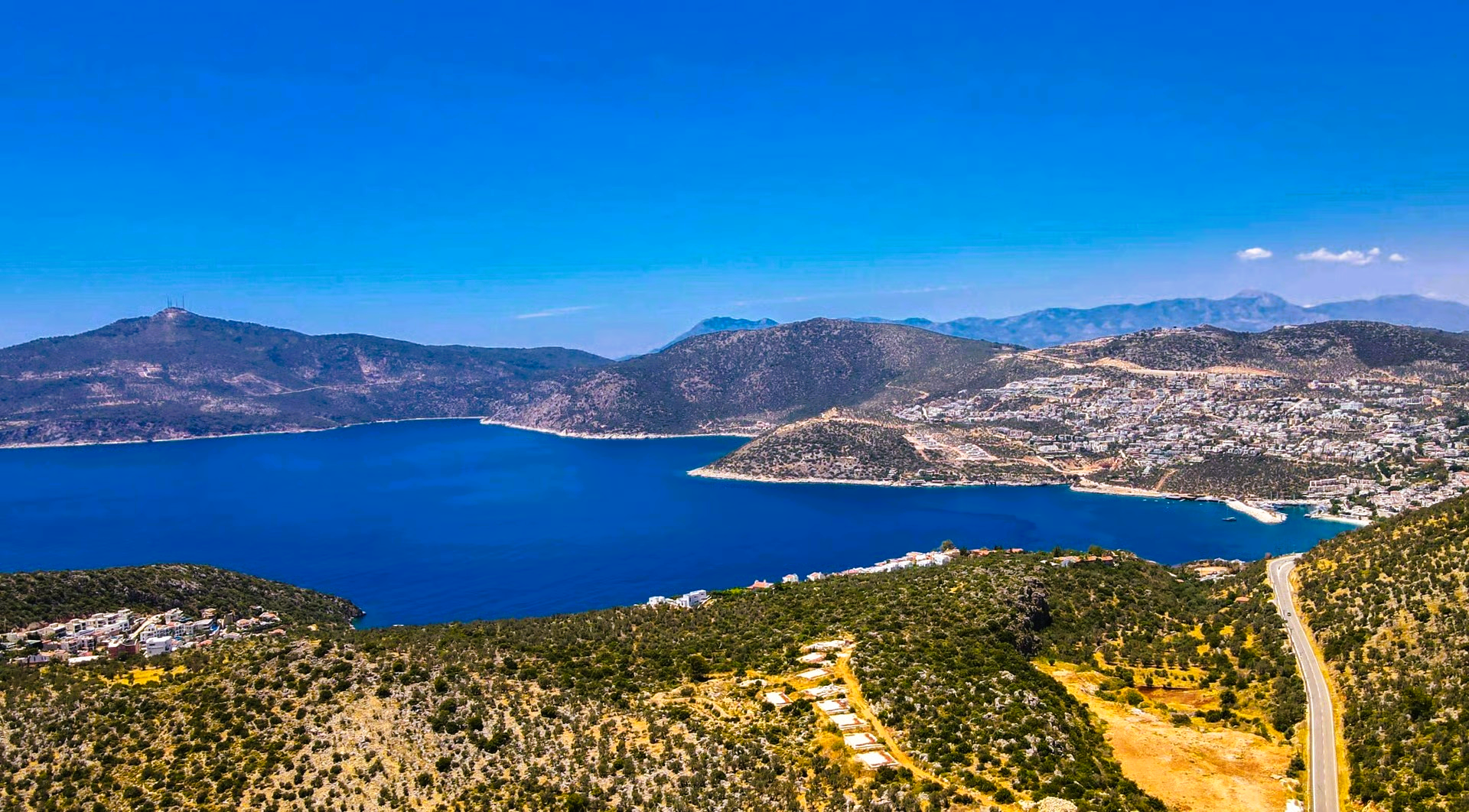 Aerial view of turquoise water and mountains under a clear blue sky, with a road winding along the coast.