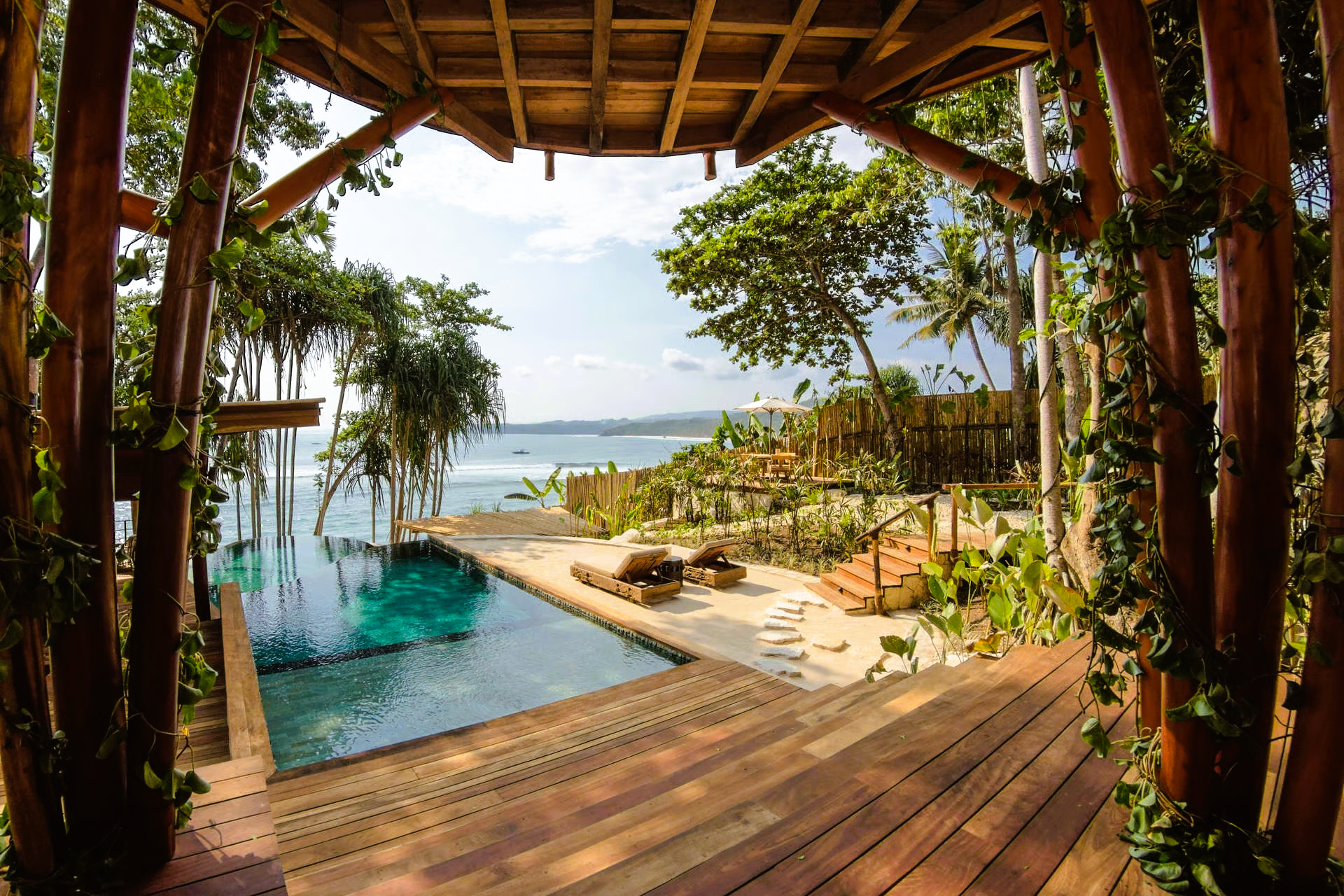 Wooden deck overlooking a turquoise pool and ocean, surrounded by trees and lush greenery.