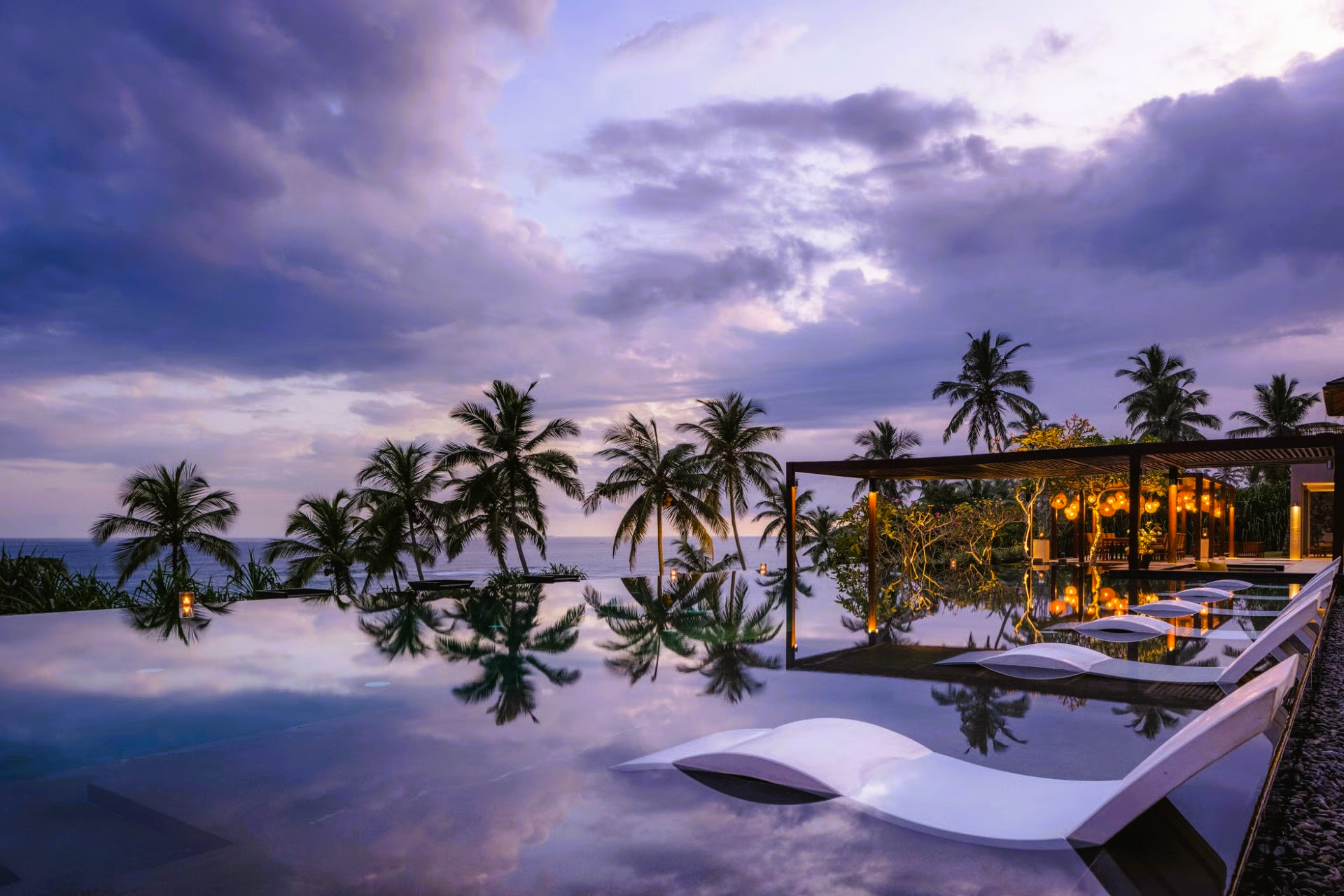 Infinity pool overlooking ocean at dusk; palm trees, white lounge chairs, dramatic sky.