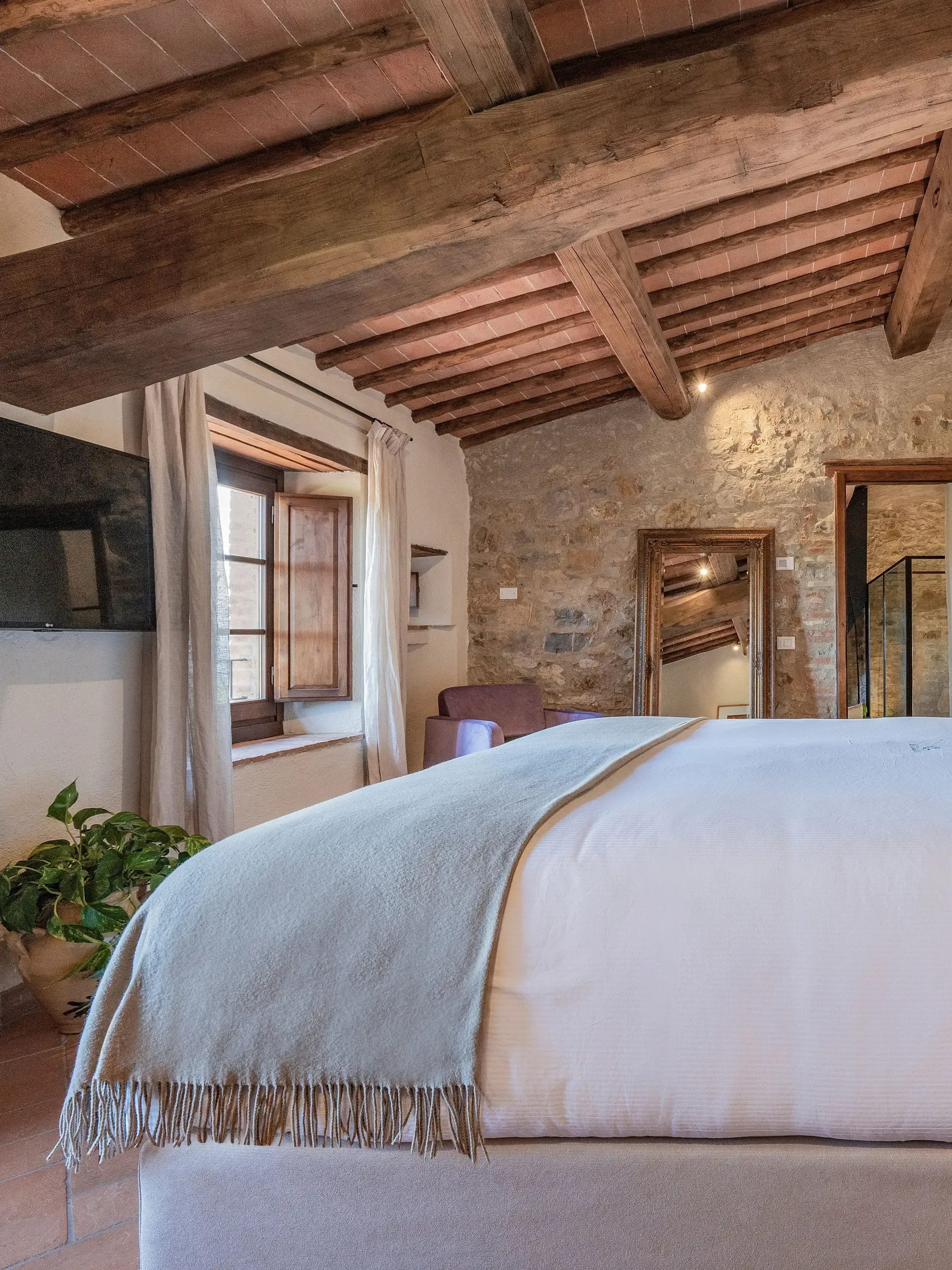 Bedroom interior with bed, exposed wood beams, stone walls, window, and mirror.