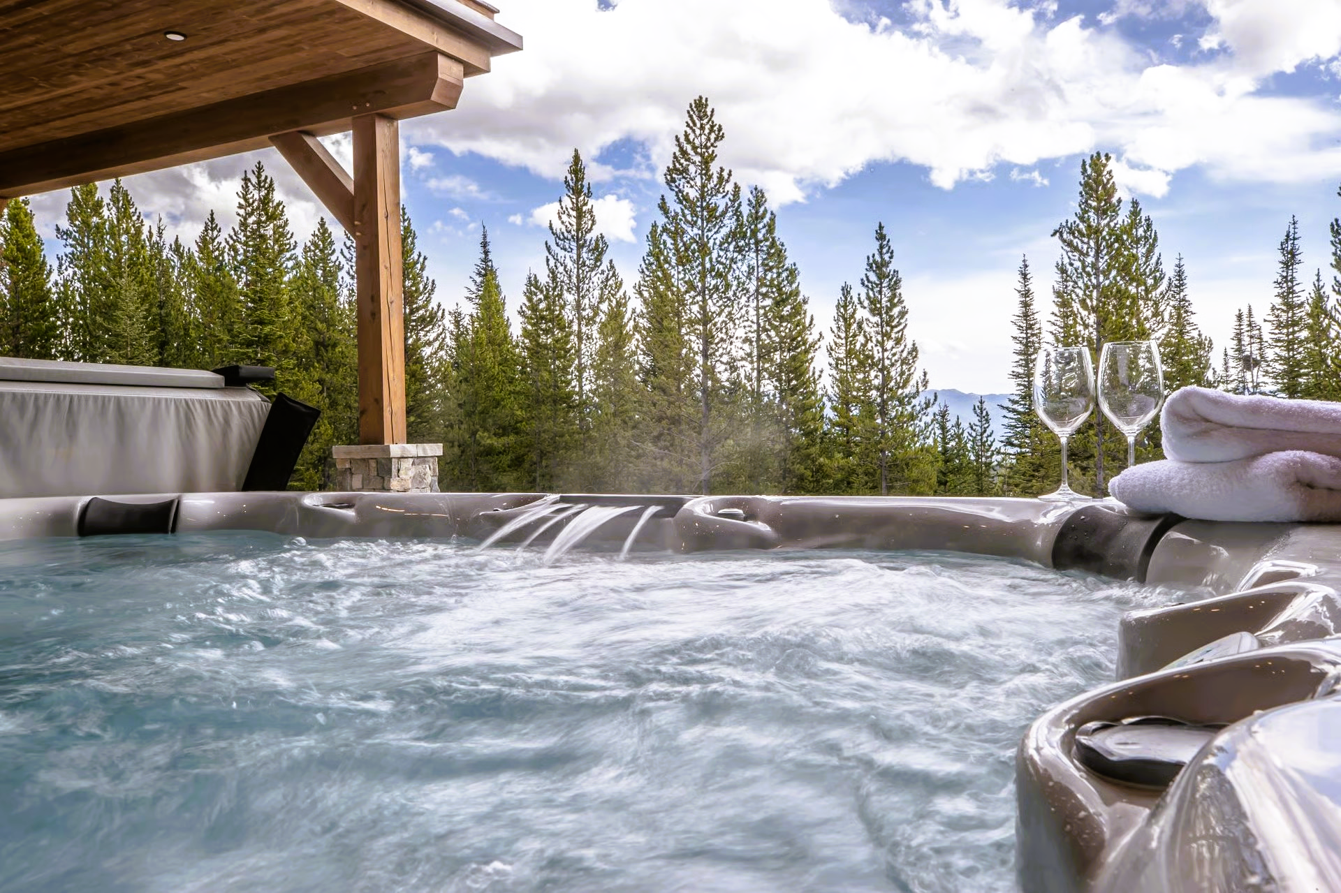 Hot tub overlooking a forest and mountains. Champagne glasses and folded towels sit nearby.