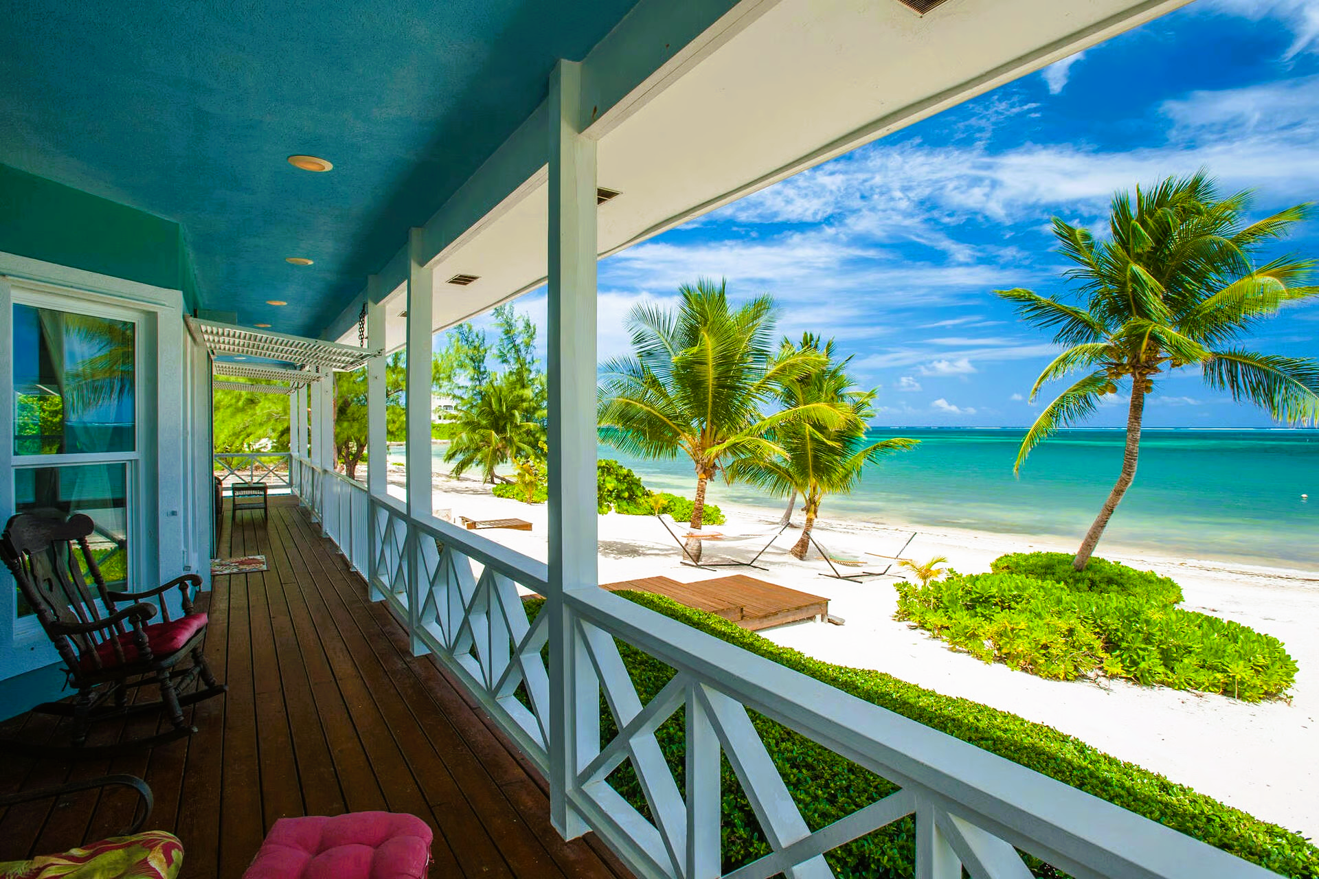 A beachfront porch with white railings, rocking chair, and view of the ocean with palm trees.