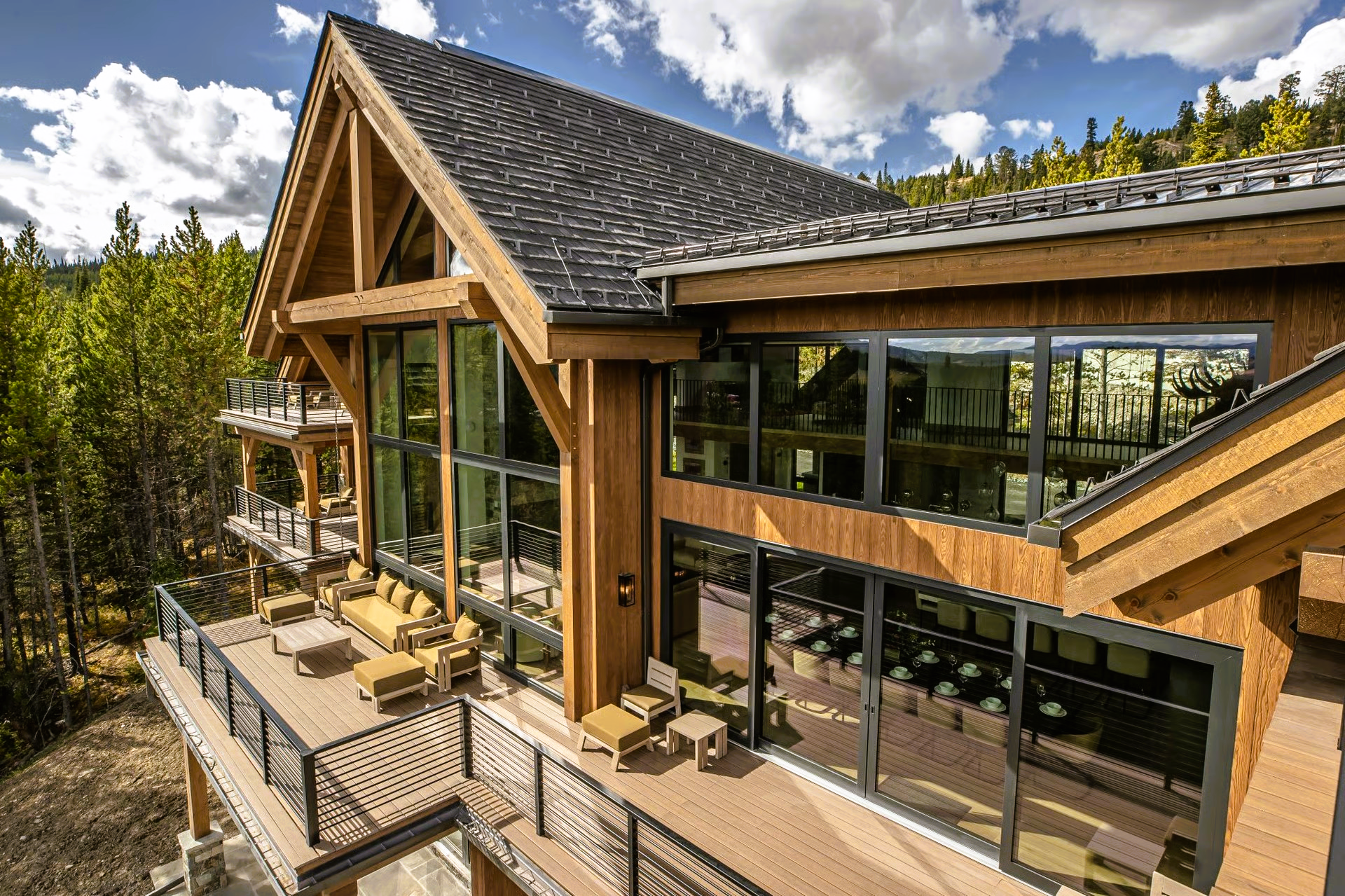 Wooden cabin with large windows and a wraparound deck overlooking a forest.