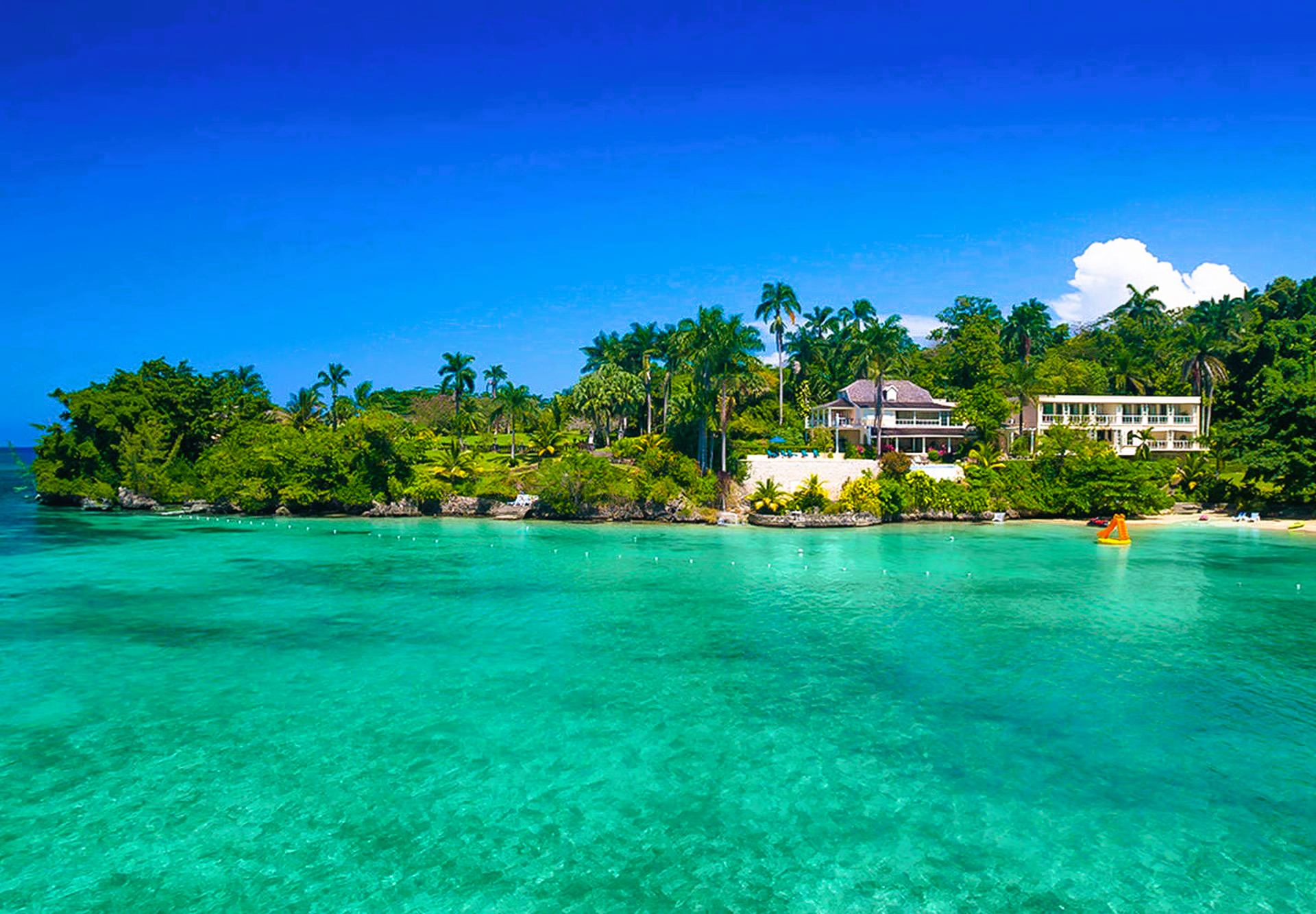 Turquoise water and a sandy beach with tropical foliage and buildings under a bright blue sky.