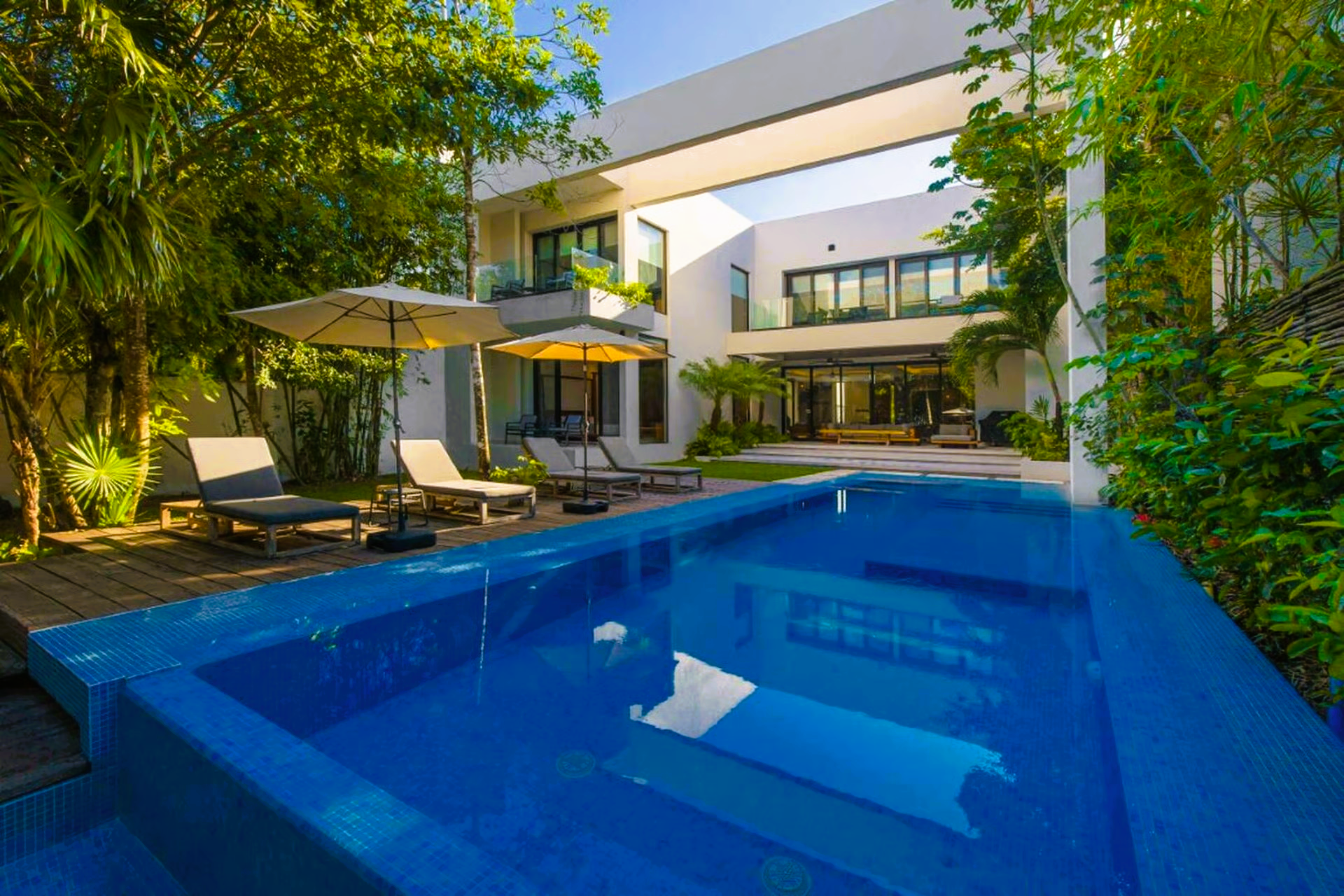 Pool with lounge chairs in front of a modern white house, surrounded by greenery. Sunny day.