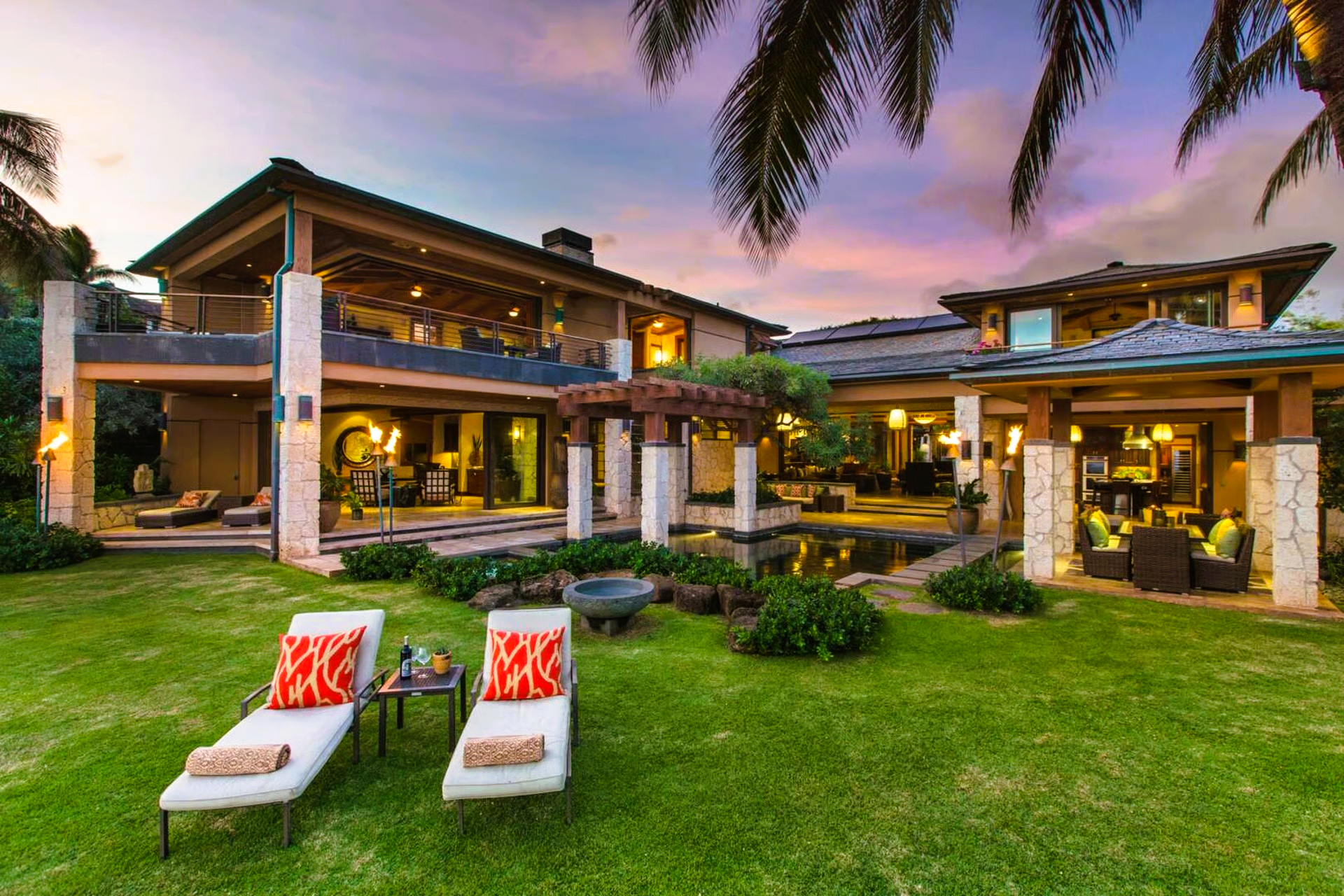 Lawn chairs on green grass face a luxury home with a pool at sunset.