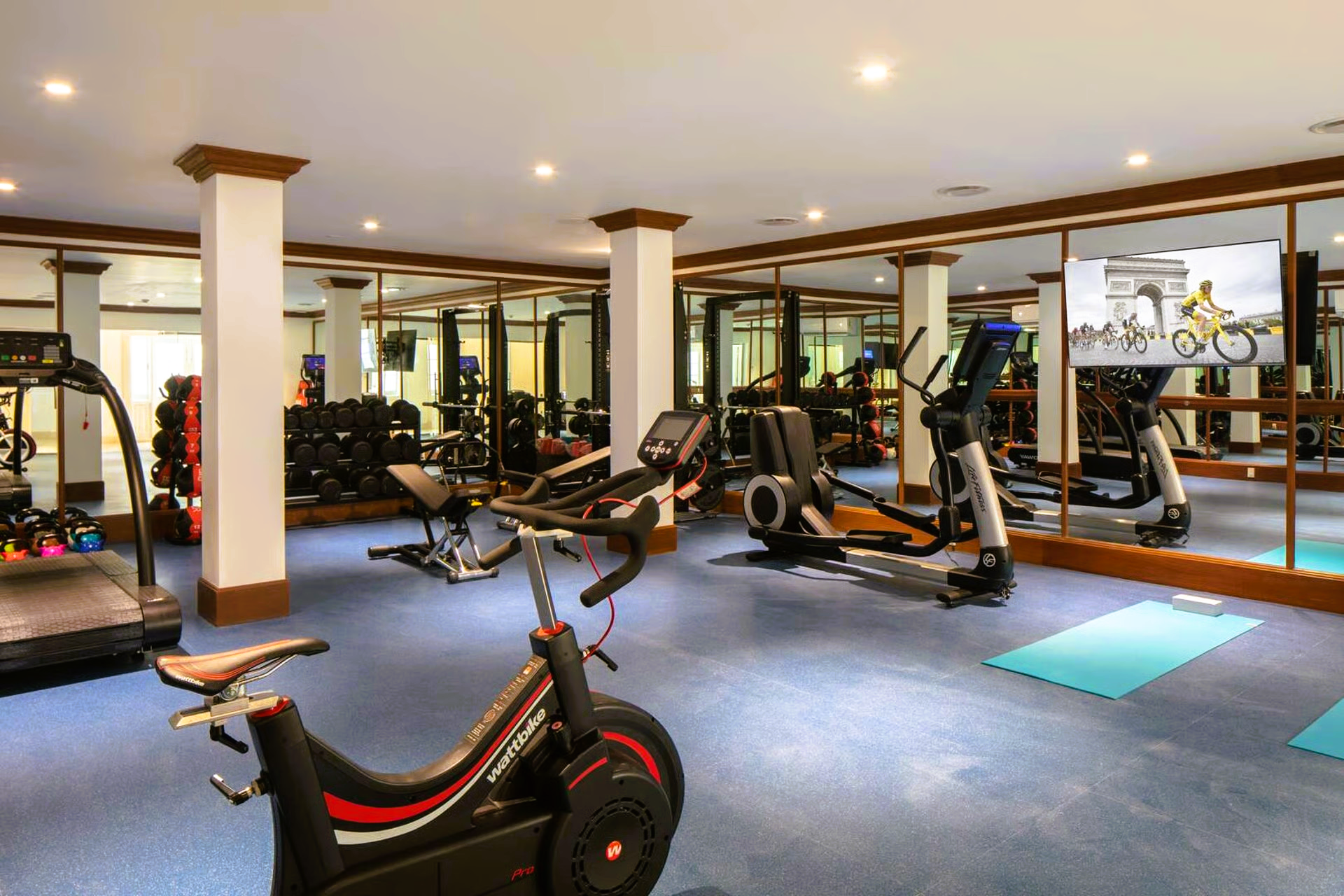 Gym with exercise machines and weights, mirrors on walls, blue carpet, and bright lighting.