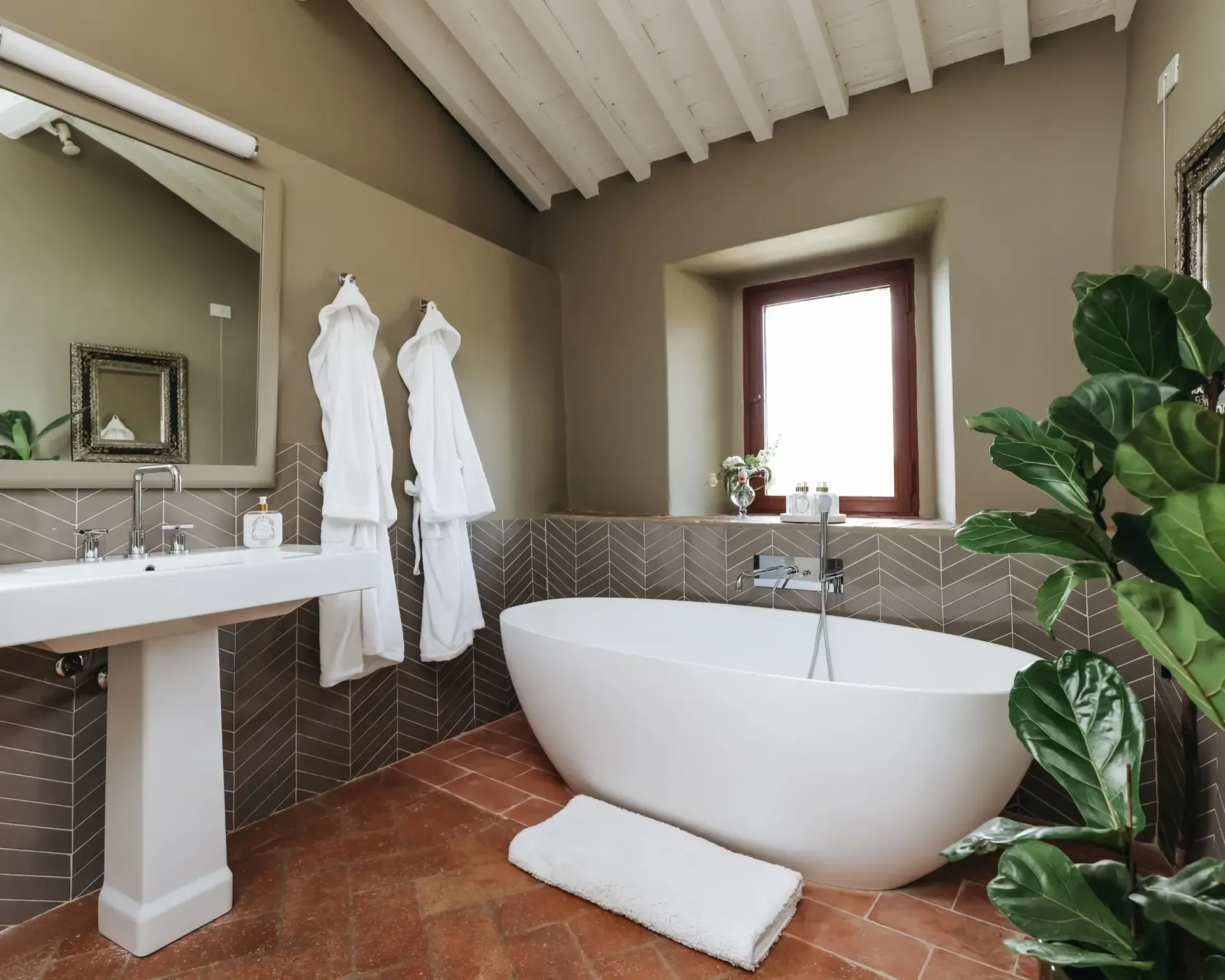 Bathroom with a freestanding white tub, pedestal sink, and window; warm tones, indoor plant.