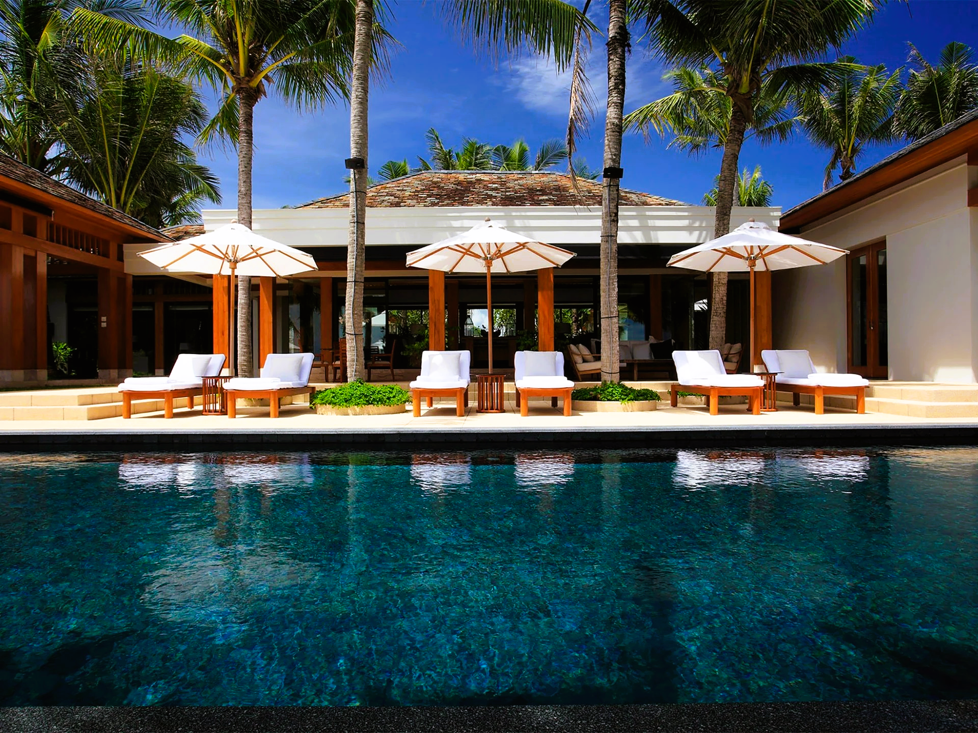 Poolside lounge chairs with umbrellas, overlooking a dark blue pool at a tropical resort.