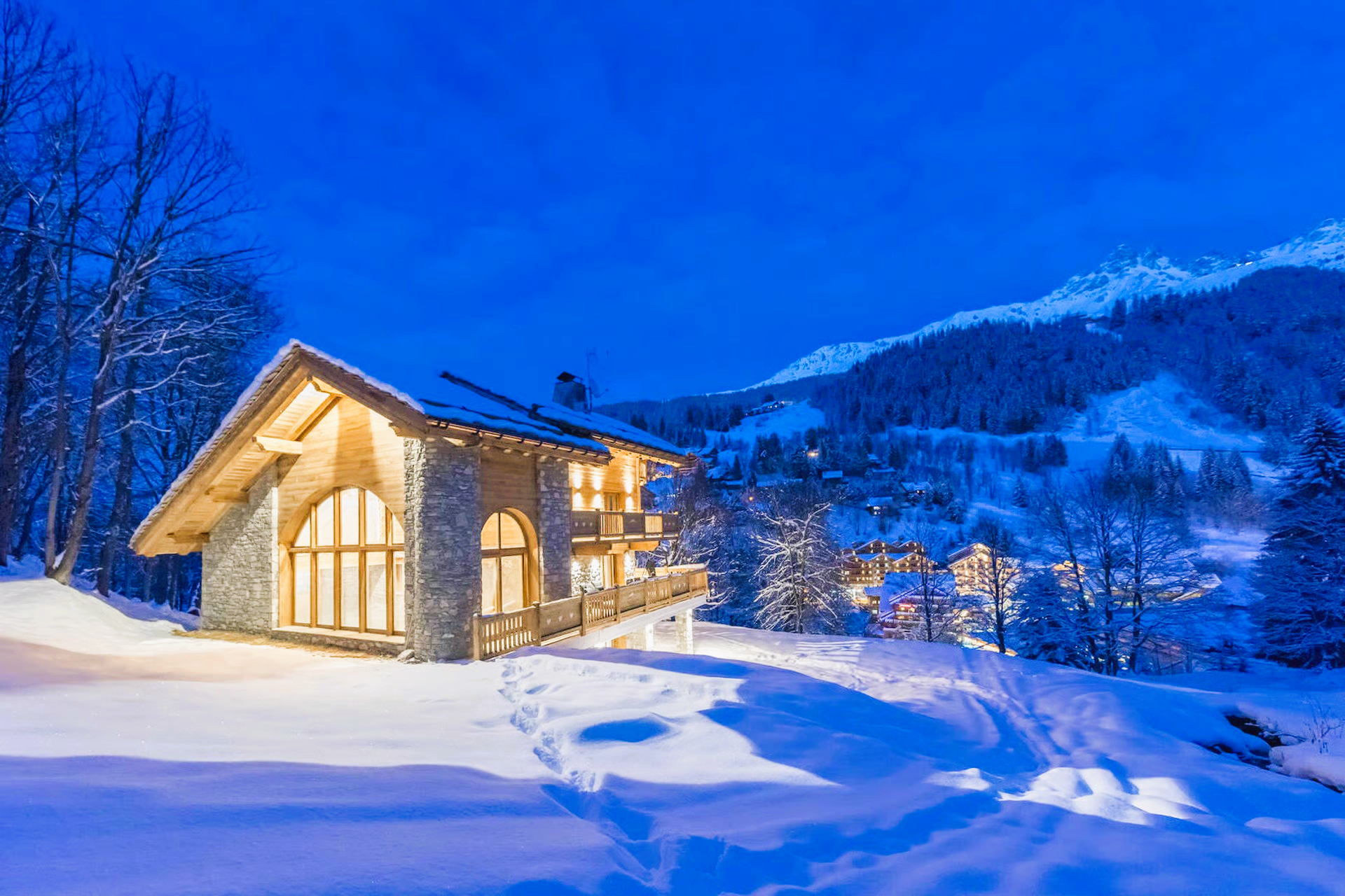 Cozy illuminated stone cabin in snowy landscape with mountains at dusk.