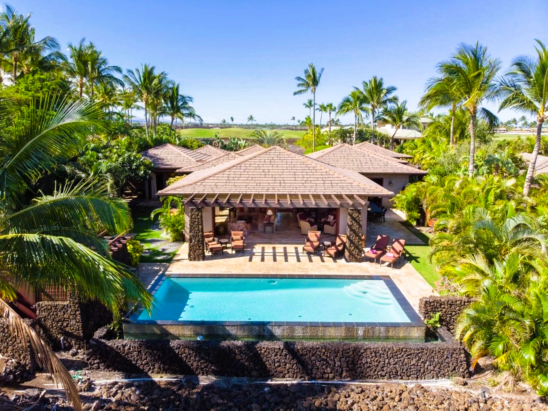 Luxurious villa with pool, wooden pergola, surrounded by lush palm trees under a clear blue sky.