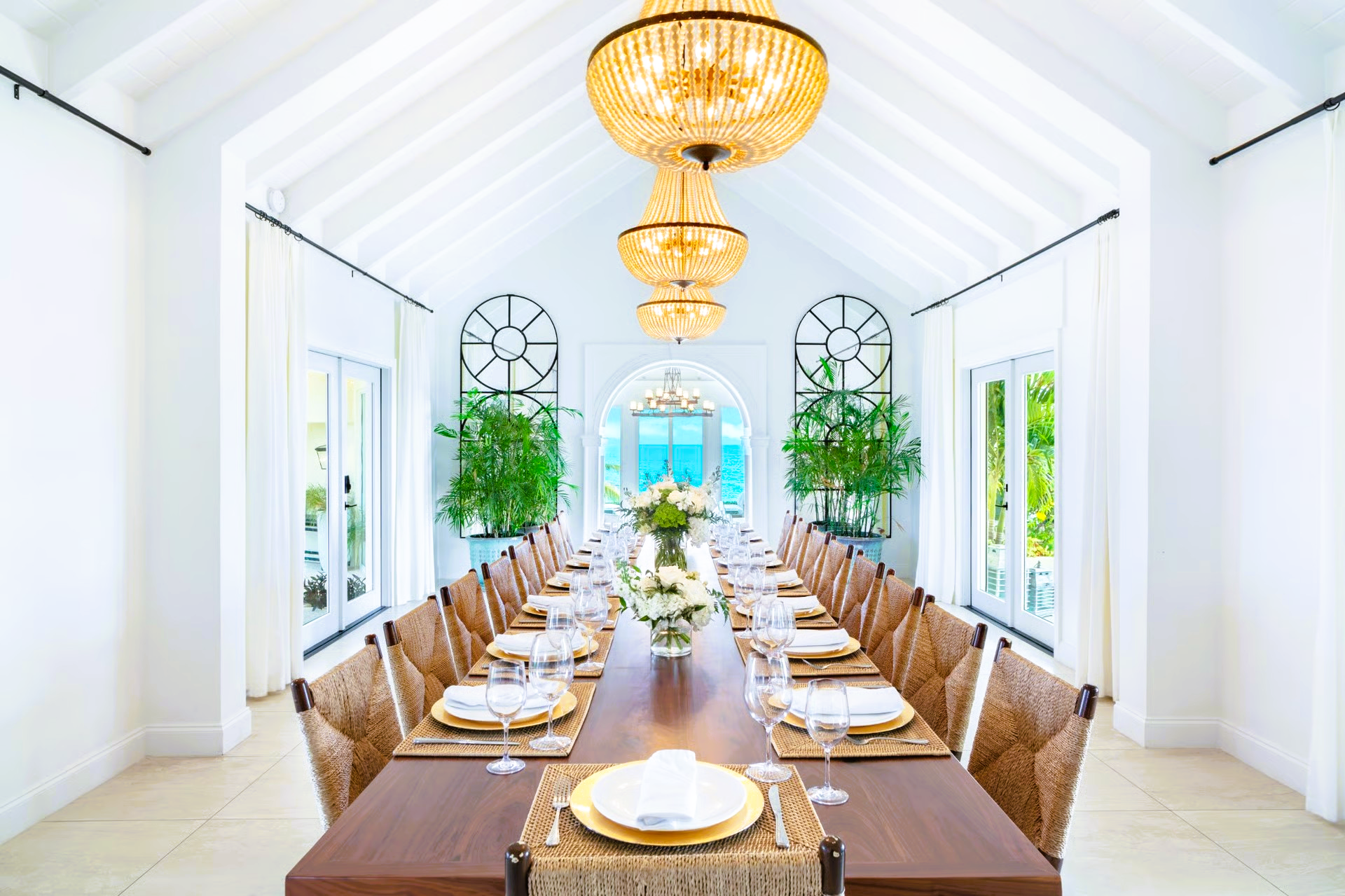 Elegant white dining room with a long table set for a large gathering, chandeliers, and ocean view.