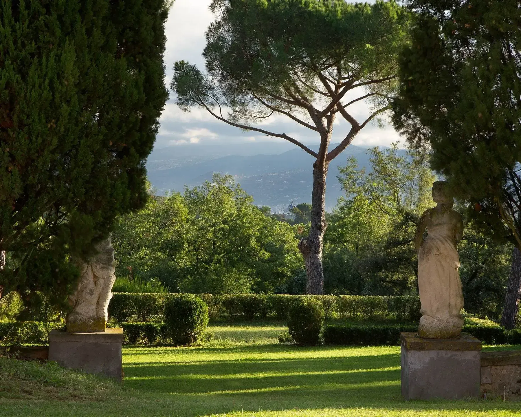 Lush Italian garden with statues, trees, and distant mountains. Sunlight filters through the leaves, casting shadows on the green grass.