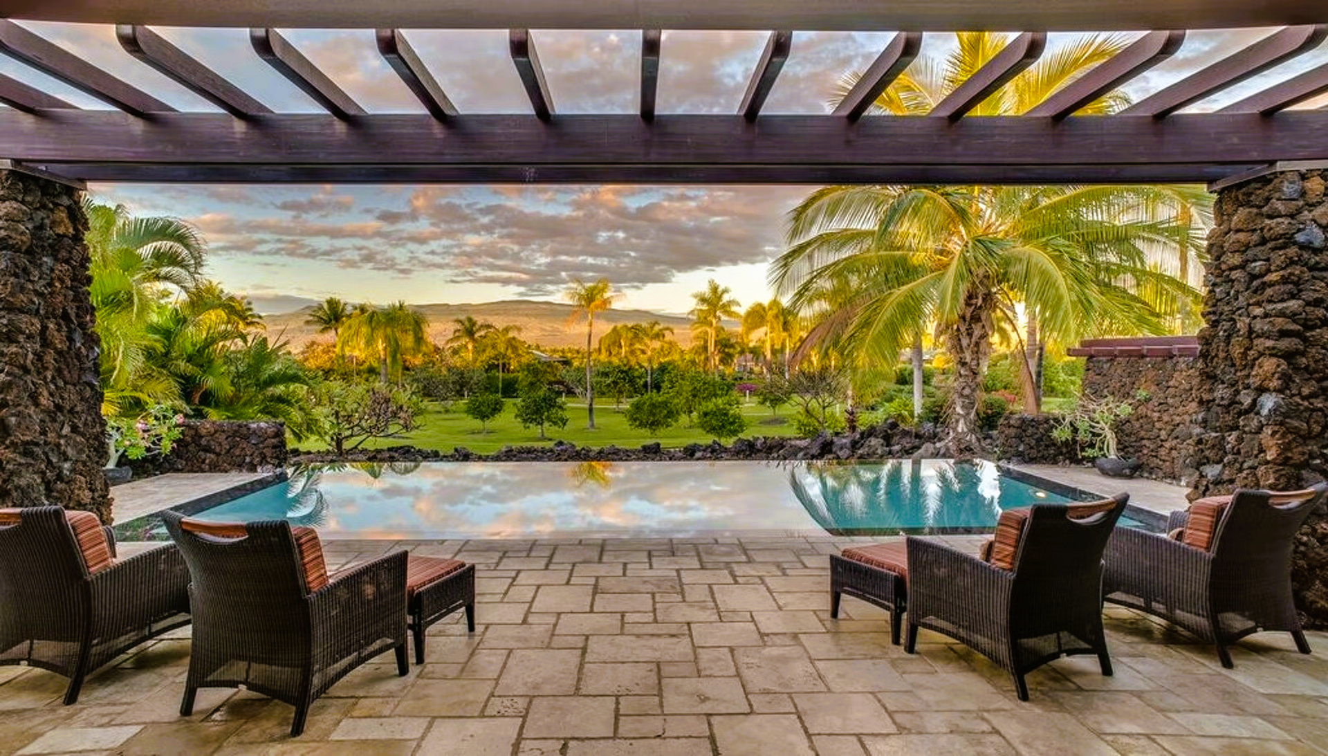 Patio with pool and mountain view, chairs, palm trees, and a sunset.