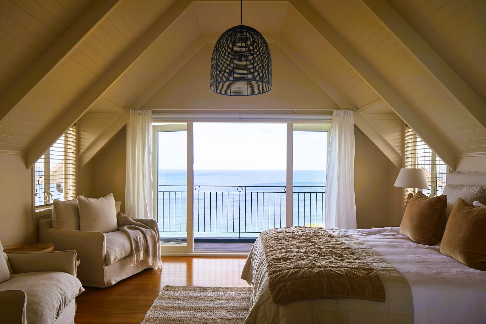 Bedroom with ocean view, neutral colors, bed, loveseat, balcony, and decorative ceiling fixture.