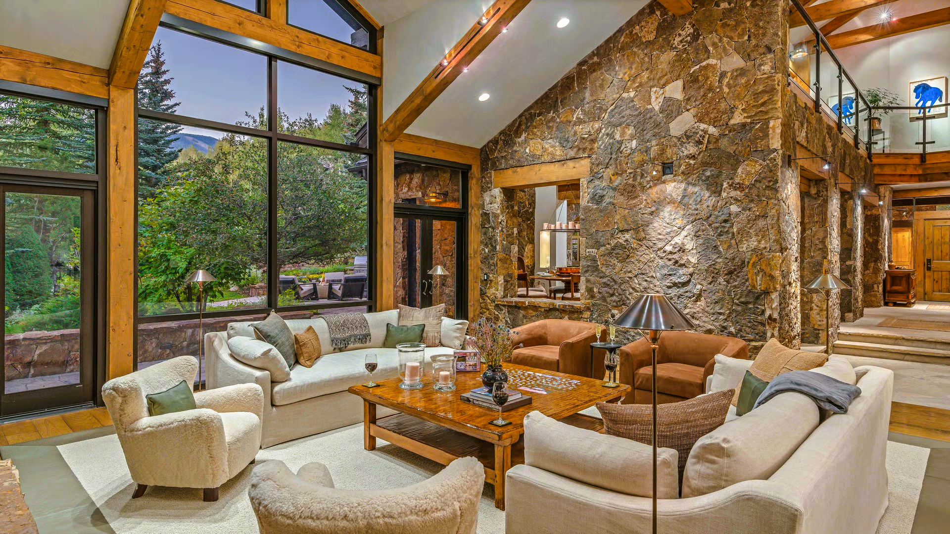 Spacious living room with floor-to-ceiling windows, stone wall, and a view of trees. Furnished with light sofas and armchairs.