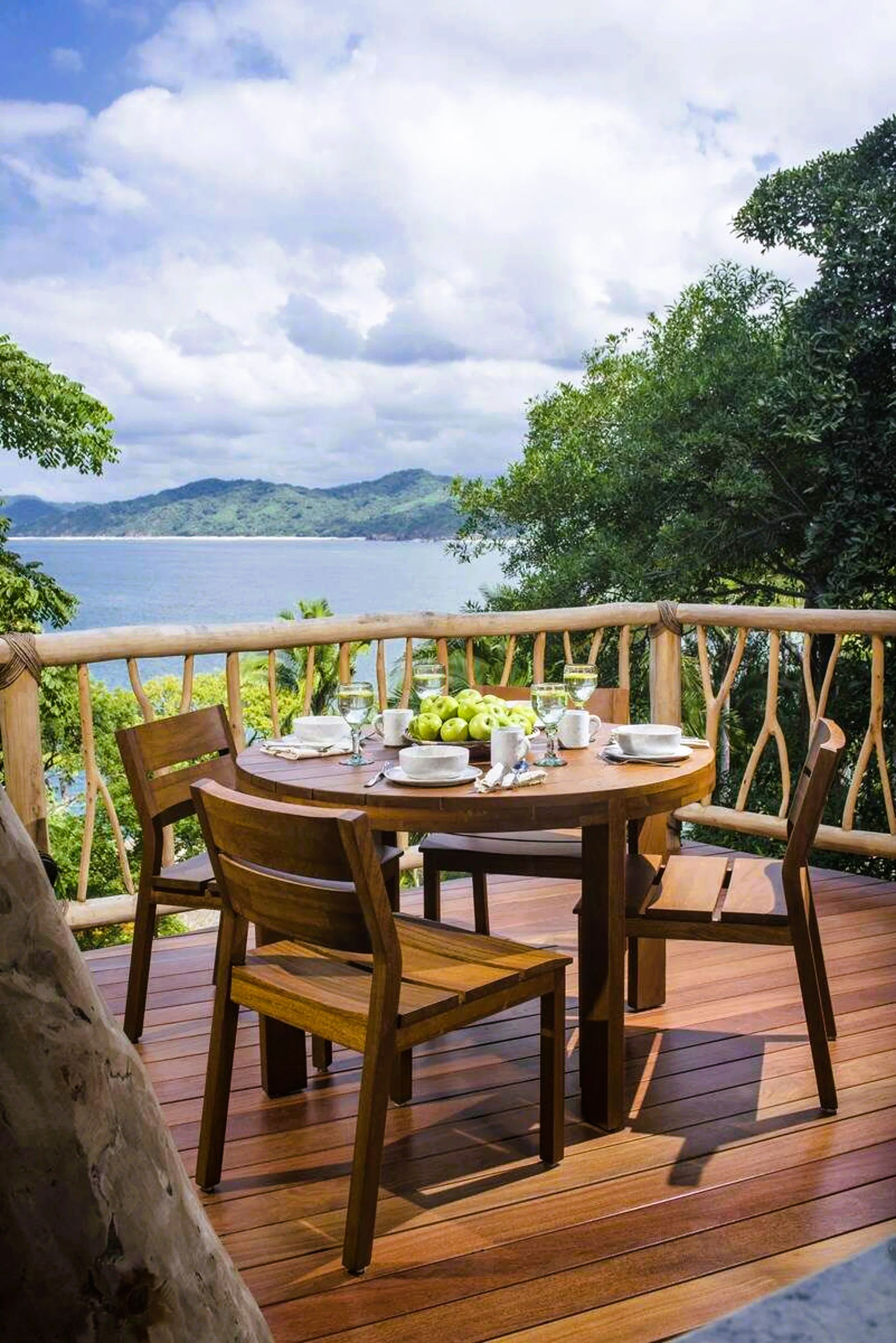 Wooden table set for a meal on a deck overlooking a bay with lush greenery and blue sky.