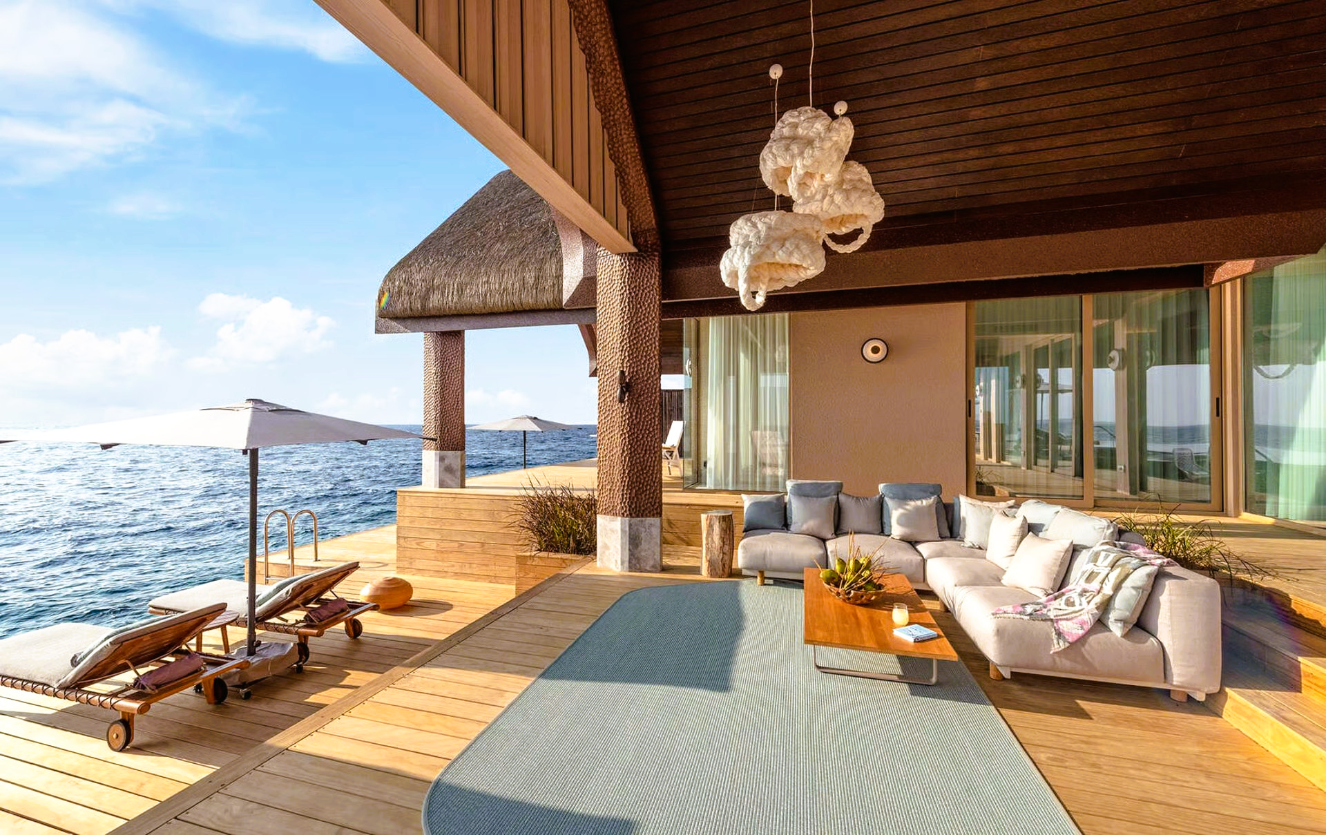 Luxury overwater villa with ocean view, lounge seating, deck, and thatched roof.