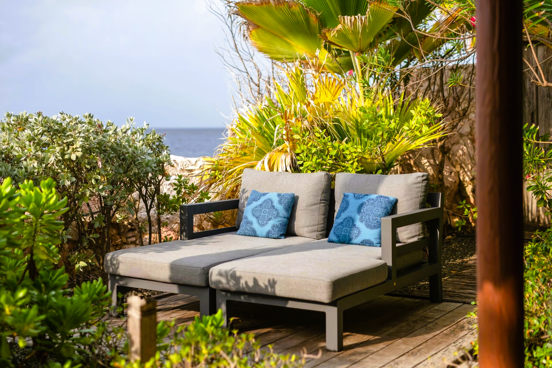 Outdoor double chaise lounge with blue pillows, near lush plants and ocean view.